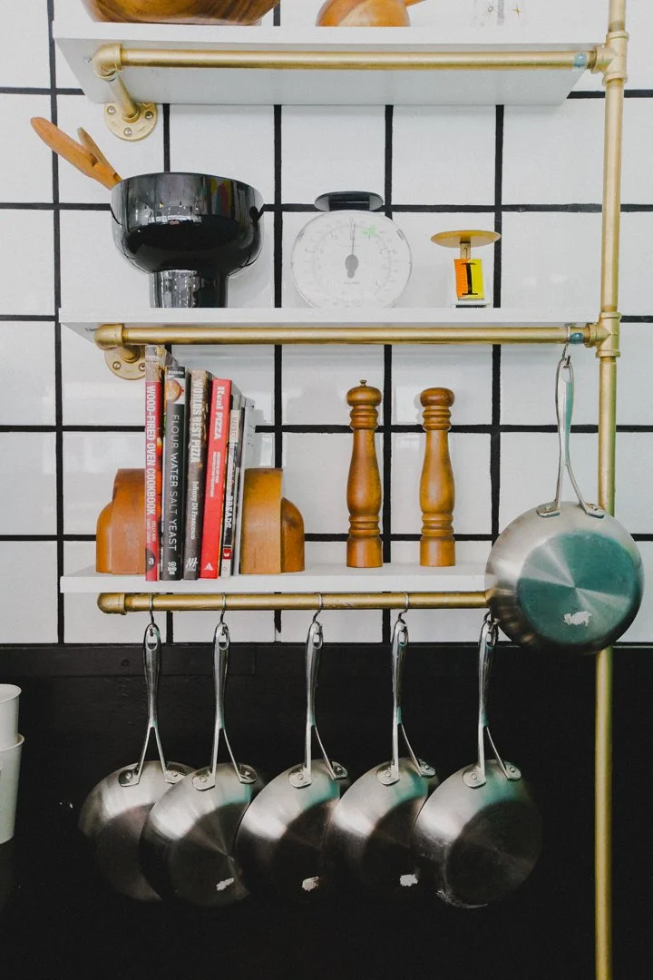 Open kitchen shelving unit with cookware, books, and kitchen tools against a white tiled wall with black grid lines.