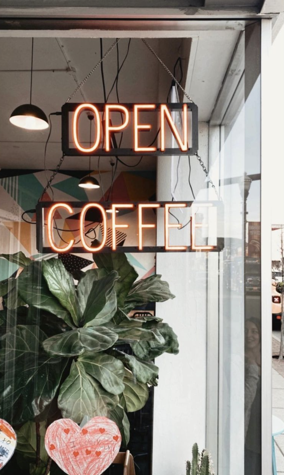 Neon signs with the words 'Open' and 'Coffee' inside a coffee shop, with plants and a window showing a person outside smiling.