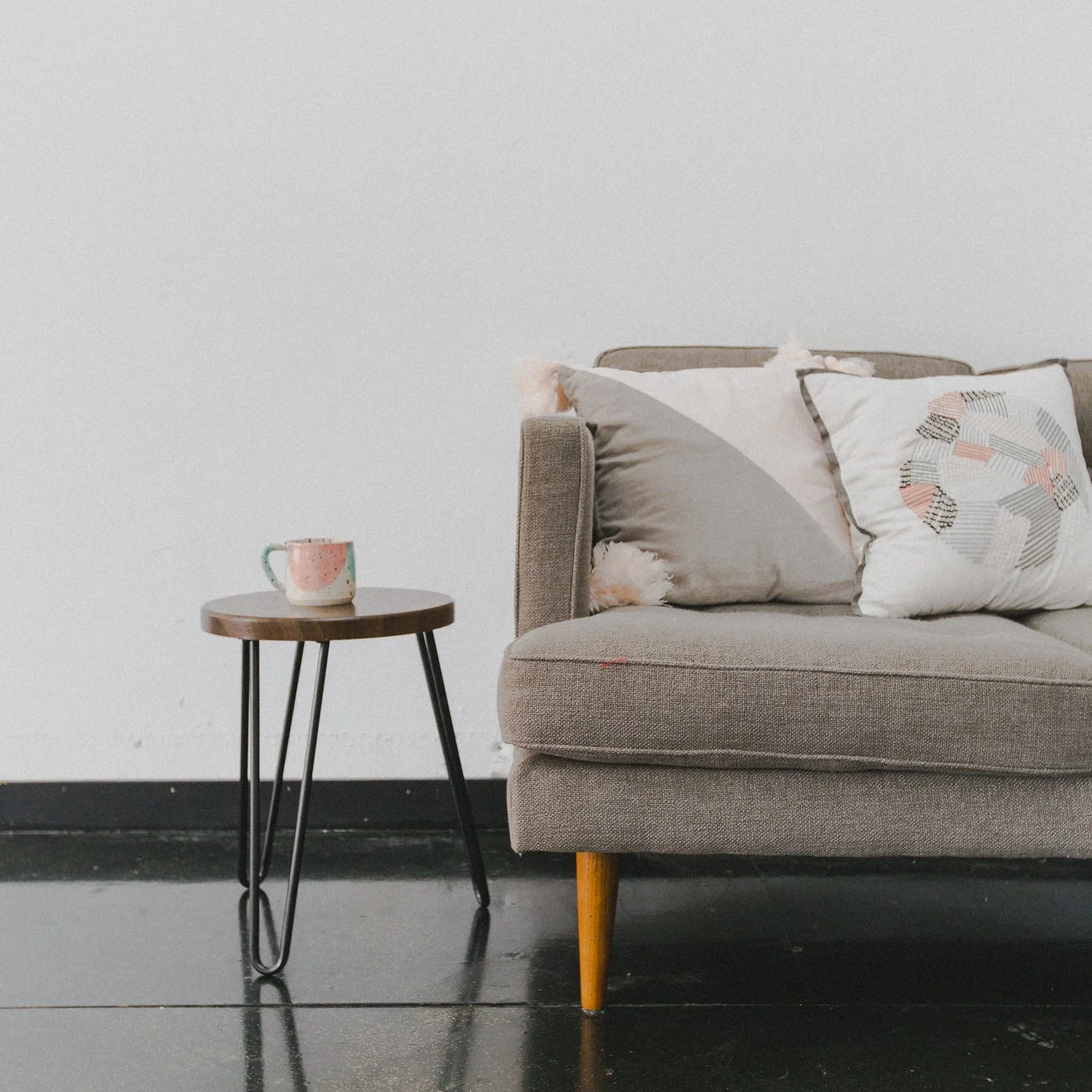 A beige sofa with pillows and a small round wooden side table with a mug on it, against a plain white wall, on a black floor.