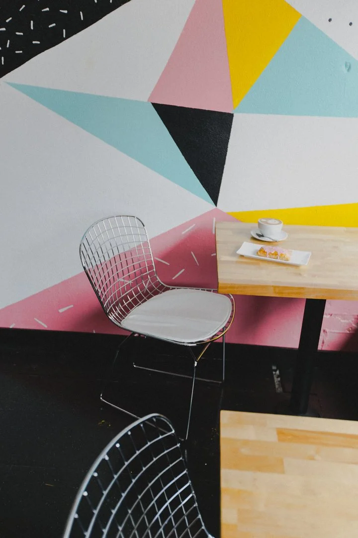 A corner of a café with a pink, black, white, light blue, and yellow geometric mural on the wall, a wooden table with a cup of coffee and a plate of small pastries, and a white wire chair.