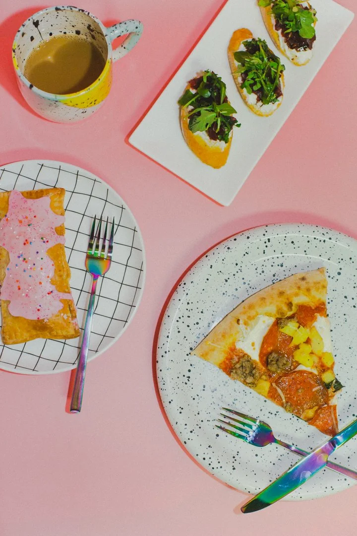 A cup of coffee, three slices of pepperoni pizza, a rectangular toast topped with pink frosting and colorful sprinkles, and three small sandwiches with leafy greens on a white platter, all on a pink surface.