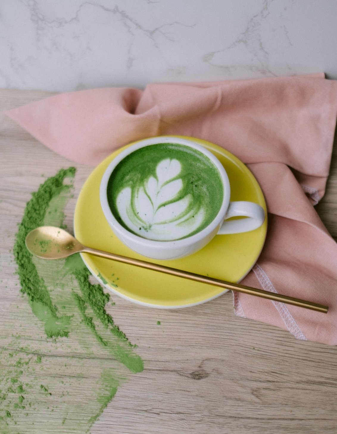A white ceramic cup of matcha latte with latte art on top, placed on a yellow saucer. Next to it, a gold spoon rests on a pink cloth napkin, with green matcha powder spilled on a light wooden surface and a marble wall in the background.