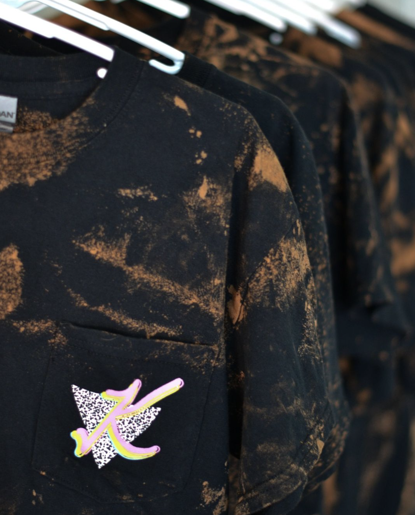 Black sweatshirt with orange dye splatter pattern and a colorful patch with a butterfly and cursive letter 'K' on the pocket, hanging on a rack.