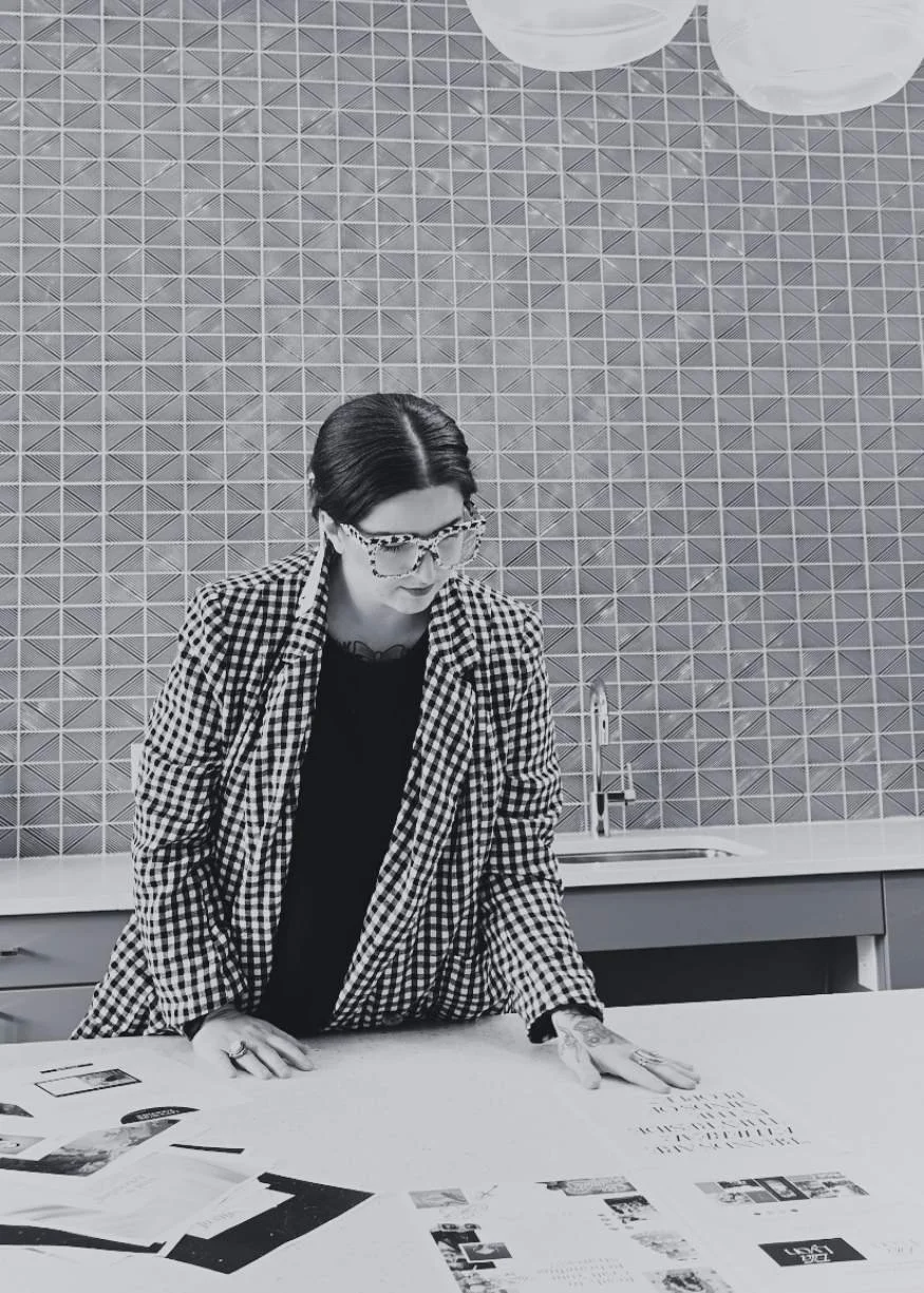 A woman with dark hair, glasses, and tattoos, wearing a checkered blazer, leaning over a table with papers and photographs, in front of a textured geometric wall.