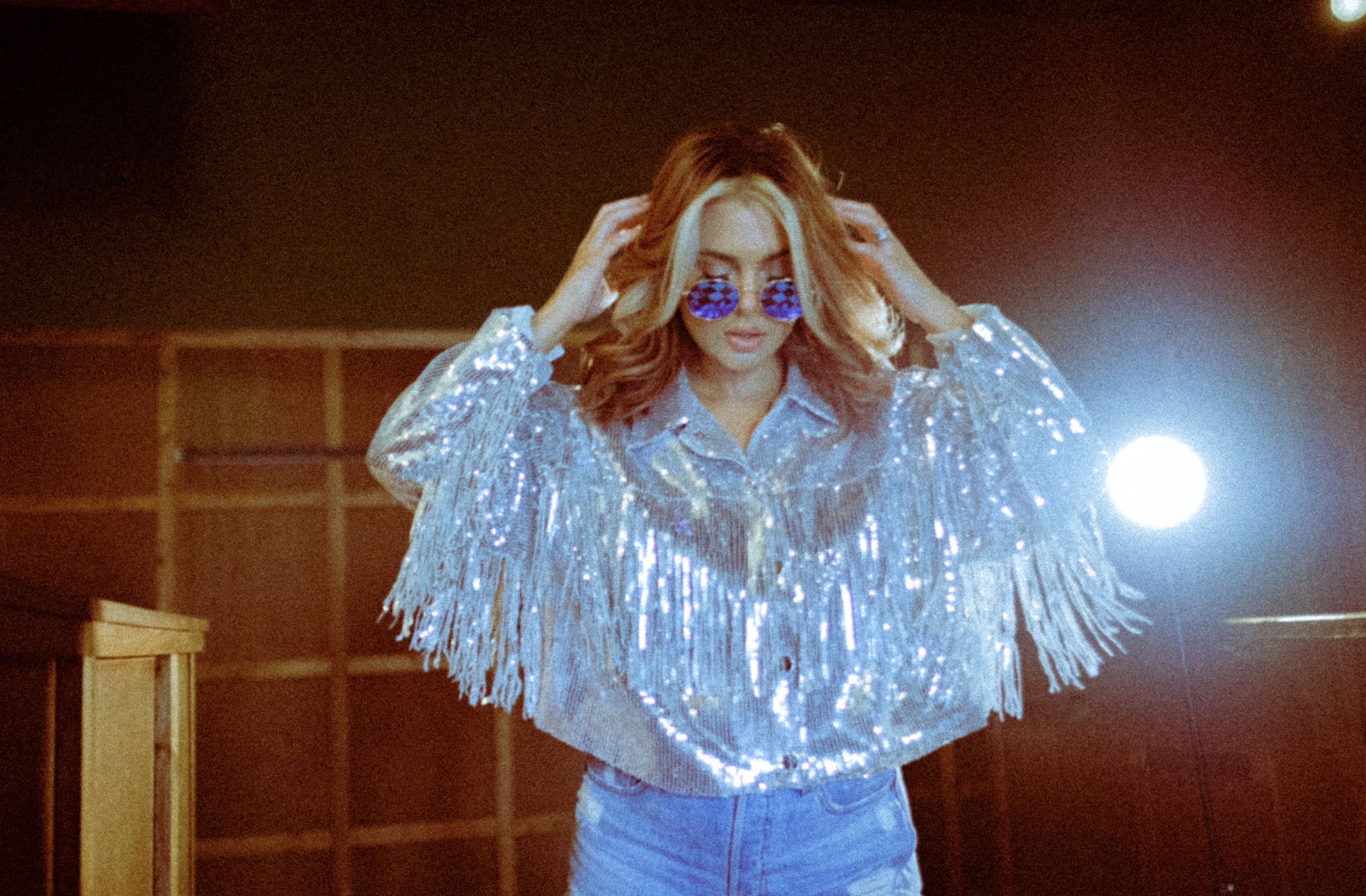 A woman with wavy red hair wearing holographic sunglasses and a shiny, fringed jacket posing indoors with a bright light behind her.