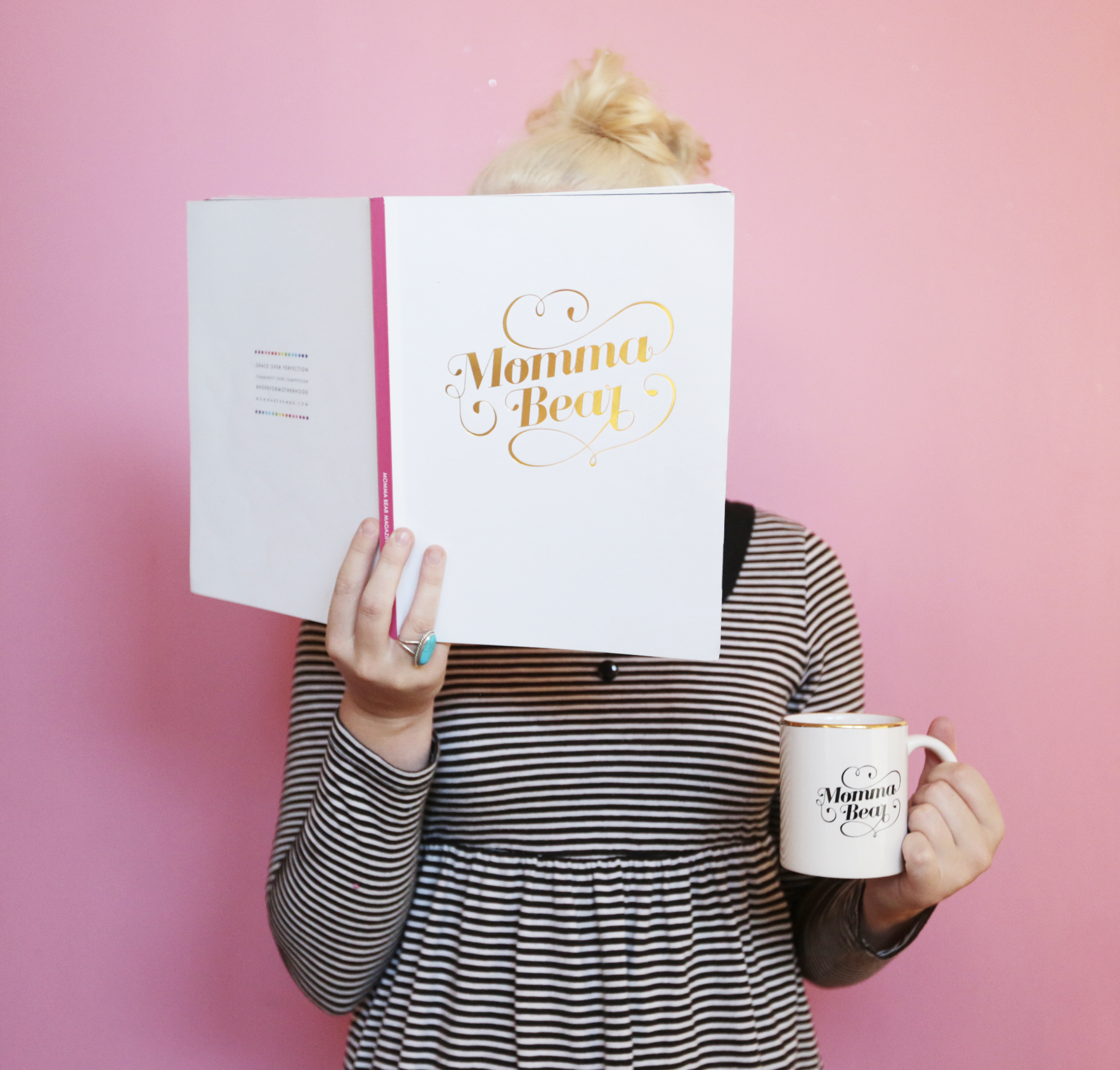 Person holding a book titled "Momma Beary" in front of their face and a mug with the same title in front of a pink background. The person is wearing a black and white striped dress and has blonde hair.