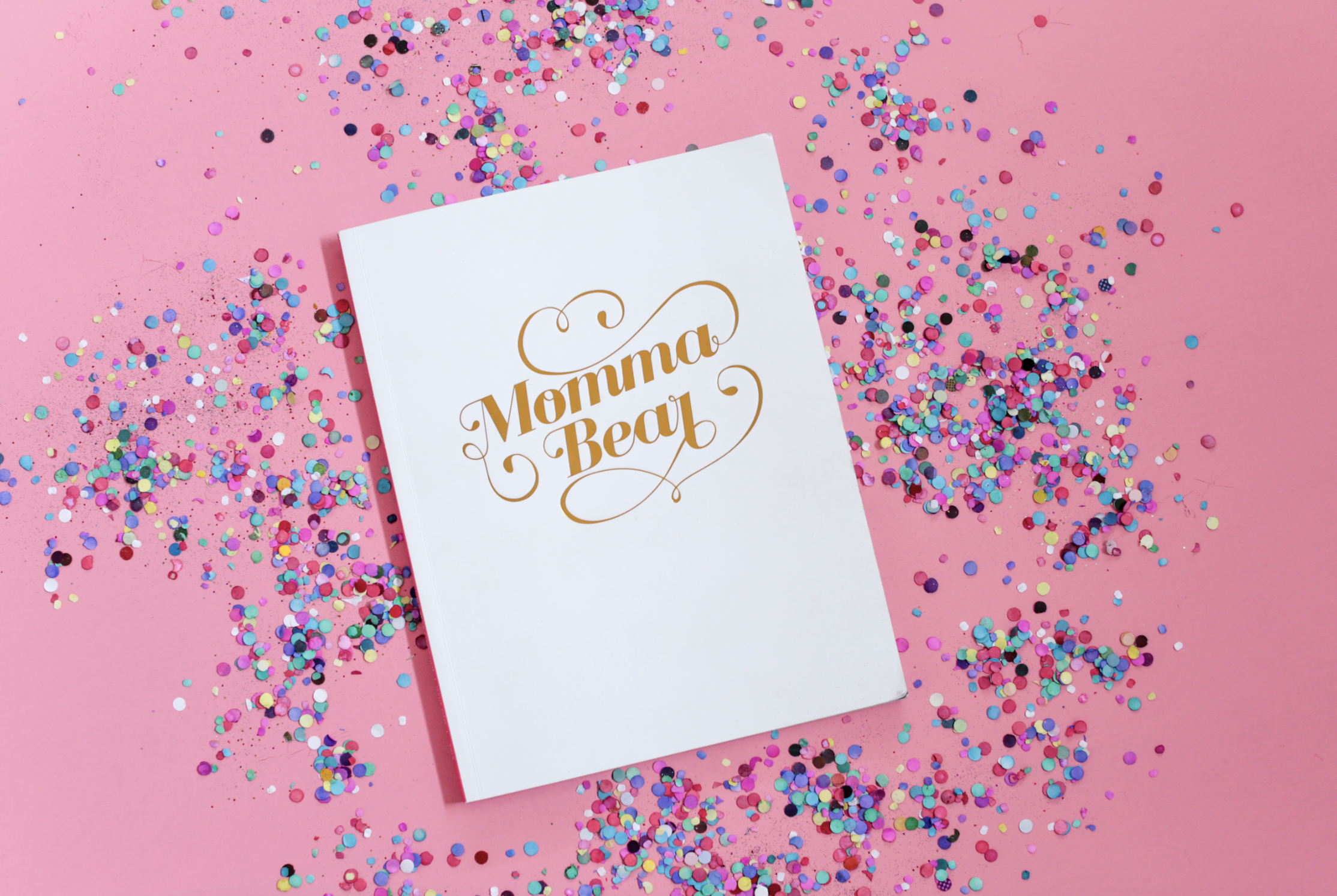 A white booklet with gold lettering that says 'Momma Bear' on a pink background, surrounded by colorful confetti.
