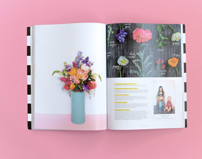 Open magazine showing a colorful flower arrangement in a turquoise vase on a white background on the left page, and a chalkboard with painted flowers and written names on the right page, set on a pink surface.