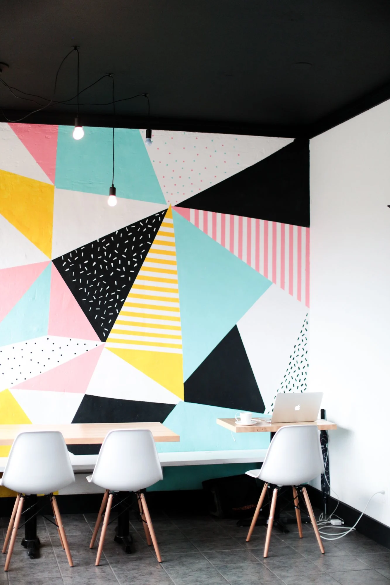 A modern workspace with a geometric mural wall, a wooden table with white chairs, a MacBook, a coffee cup, and hanging light bulbs.