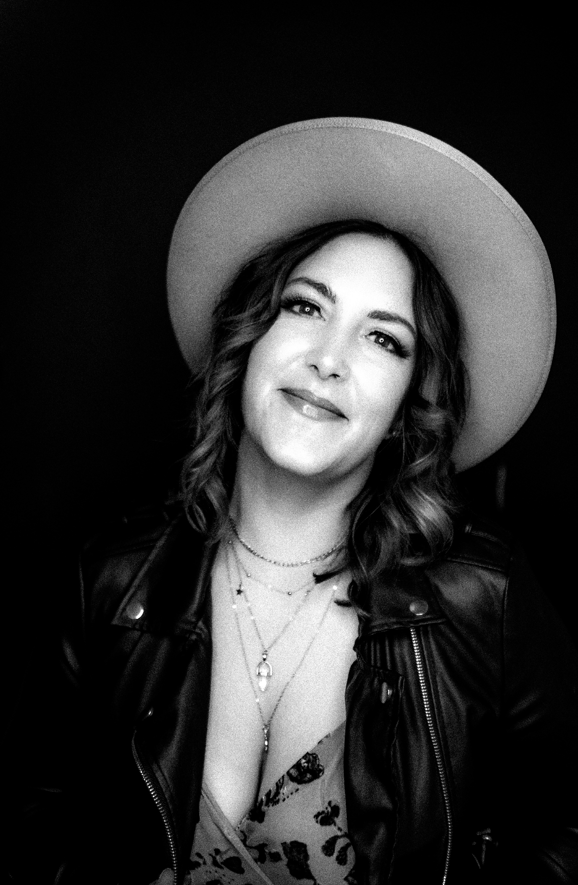 Black and white portrait of a woman wearing a wide-brimmed hat, leather jacket, layered necklaces, and a patterned top, smiling softly.