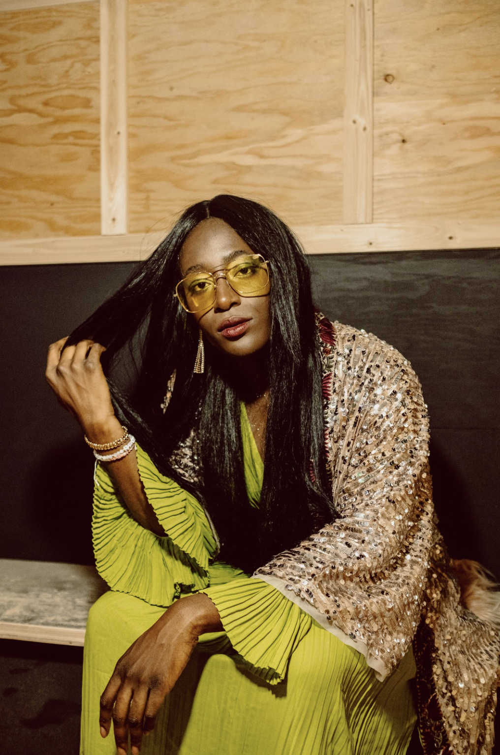 A woman with long black hair wearing yellow-tinted glasses, a shiny sequined jacket, a lime green pleated dress, and jewelry, sitting against a wood-paneled wall.