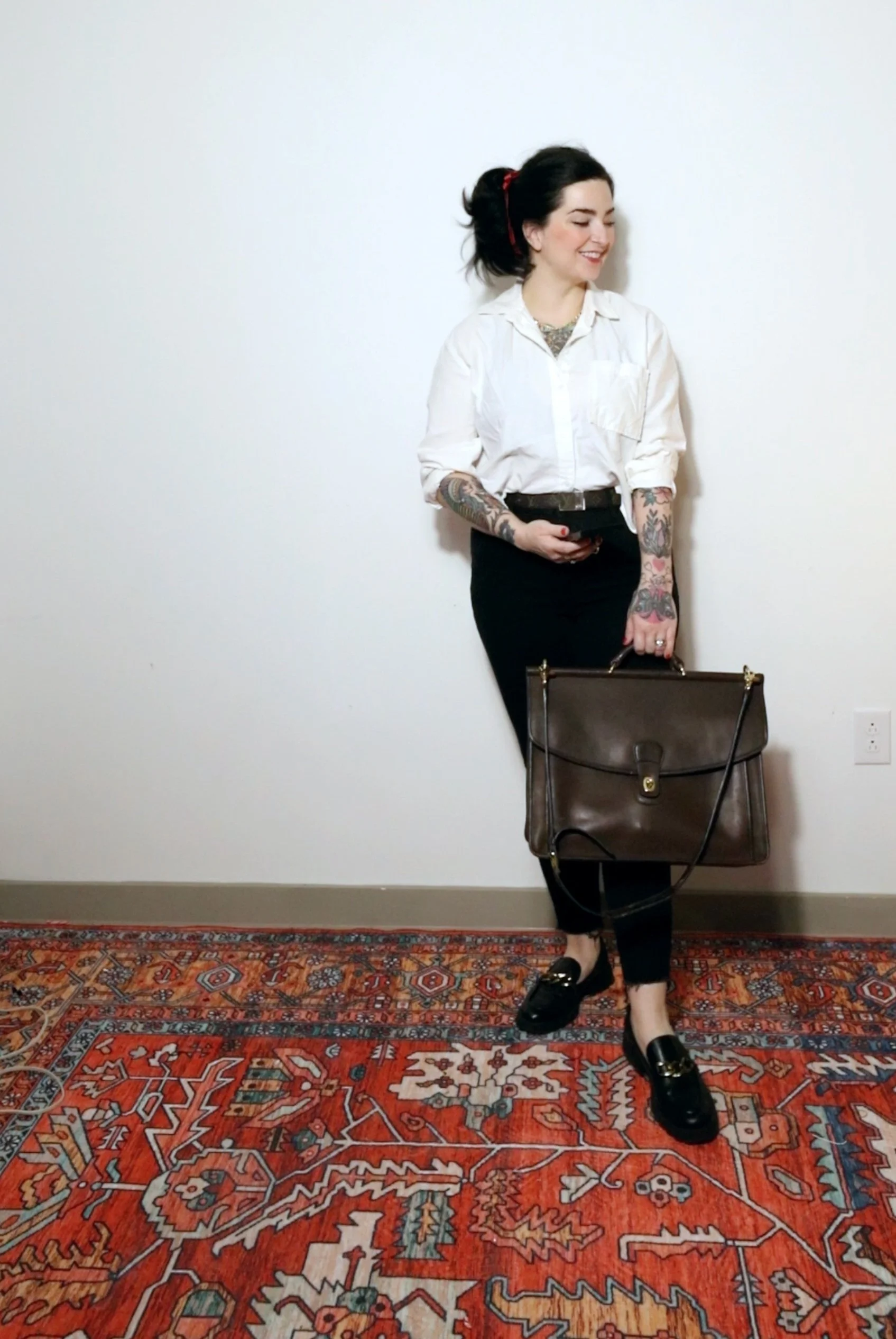 A woman with dark hair styled in an updo, wearing a white button-up shirt, black pants, and loafers, stands against a plain white wall, holding a large brown handbag and smiling.