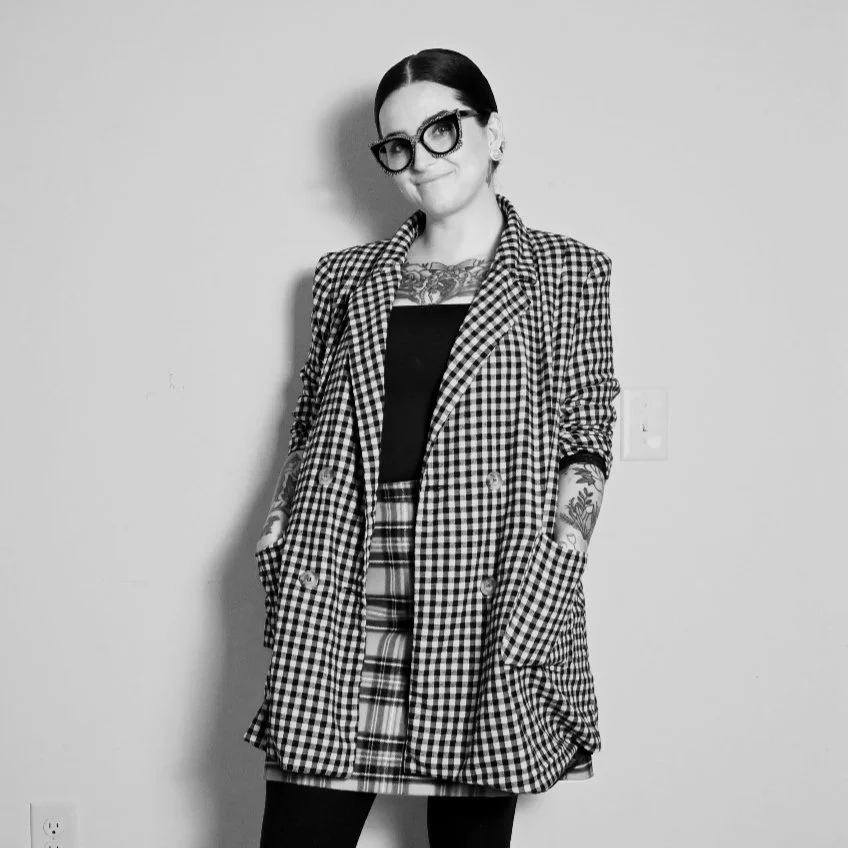 Black and white photo of a woman with dark hair, wearing large glasses, a checkered blazer, tattooed arms, and a patterned skirt, standing against a plain wall.
