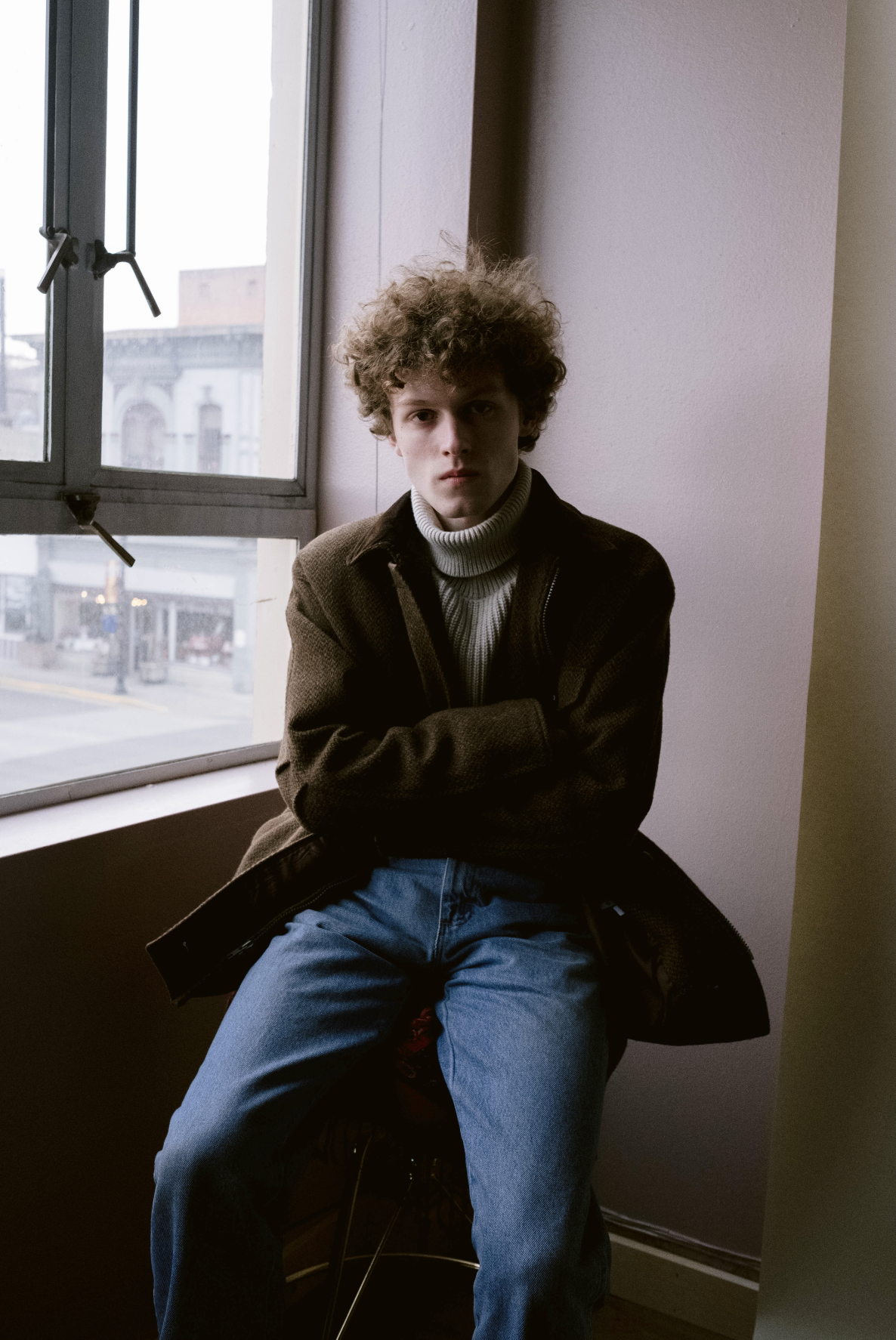 A young man with curly hair, wearing a brown jacket and a turtleneck, sitting by a window with crossed arms, looking at the camera indoors.