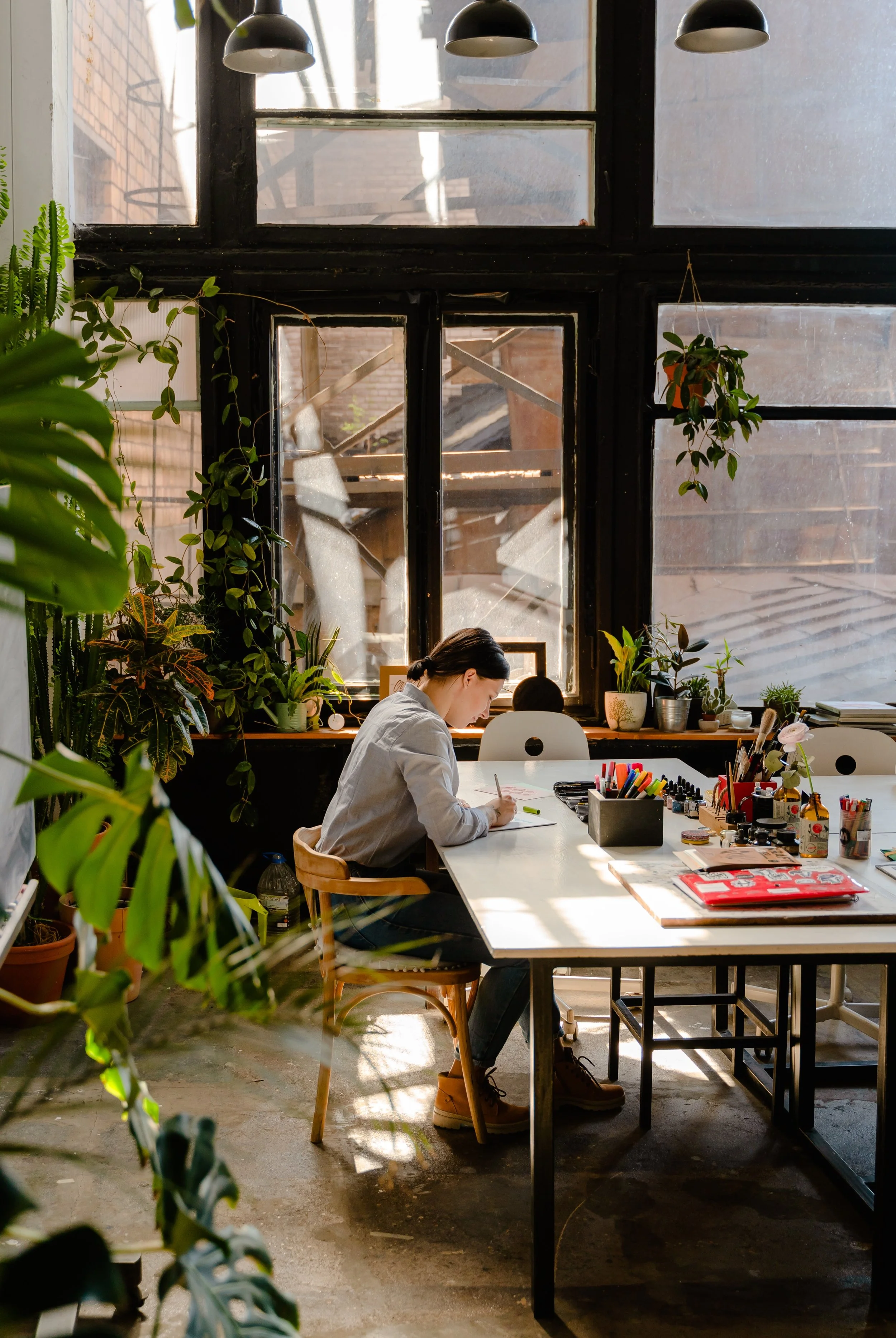 A woman working at a table in a sunlit room filled with plants and large windows.