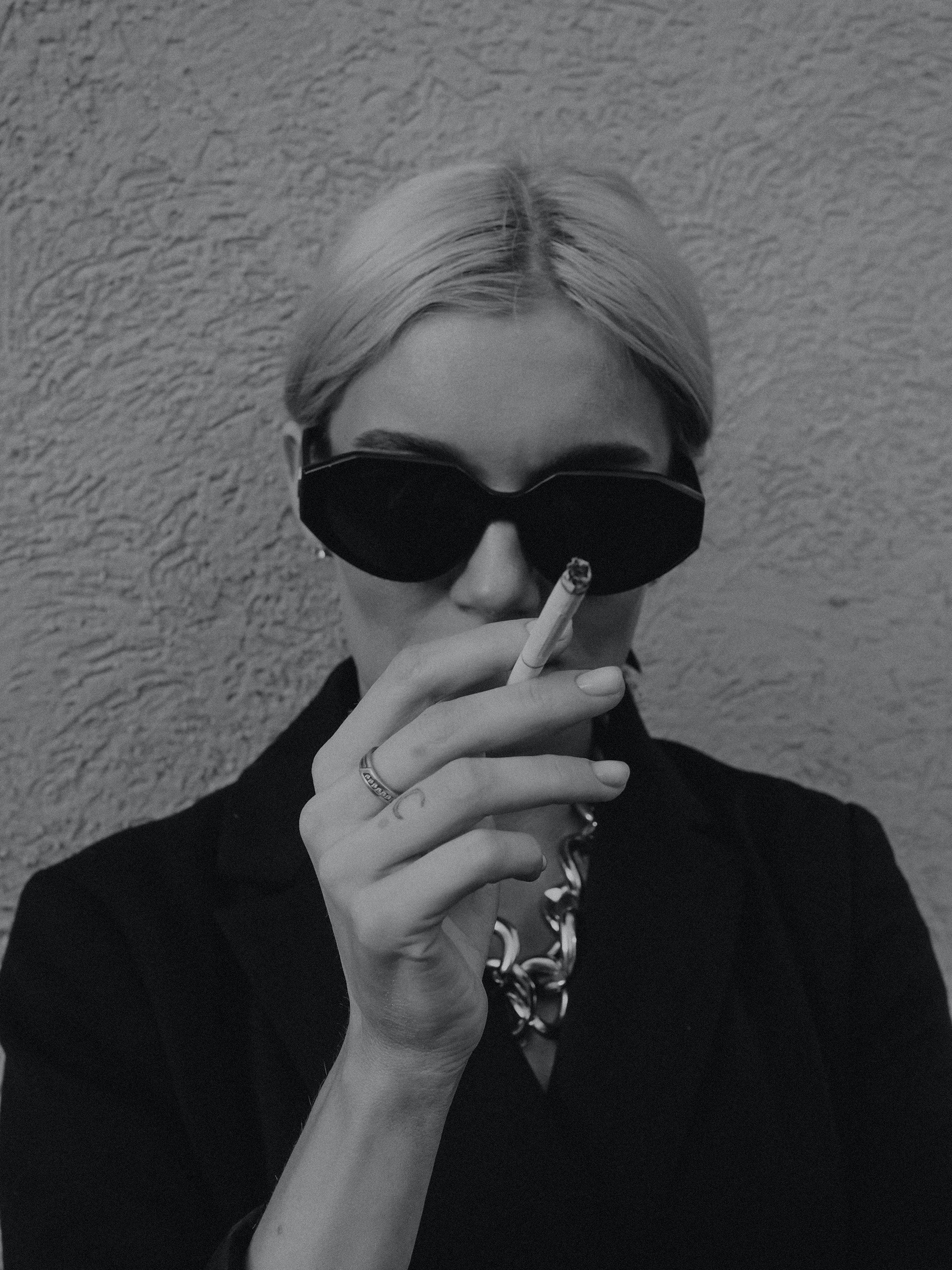 A woman with blonde hair wearing dark sunglasses and a chunky chain necklace, holding a cigarette to her lips, standing against a textured wall.