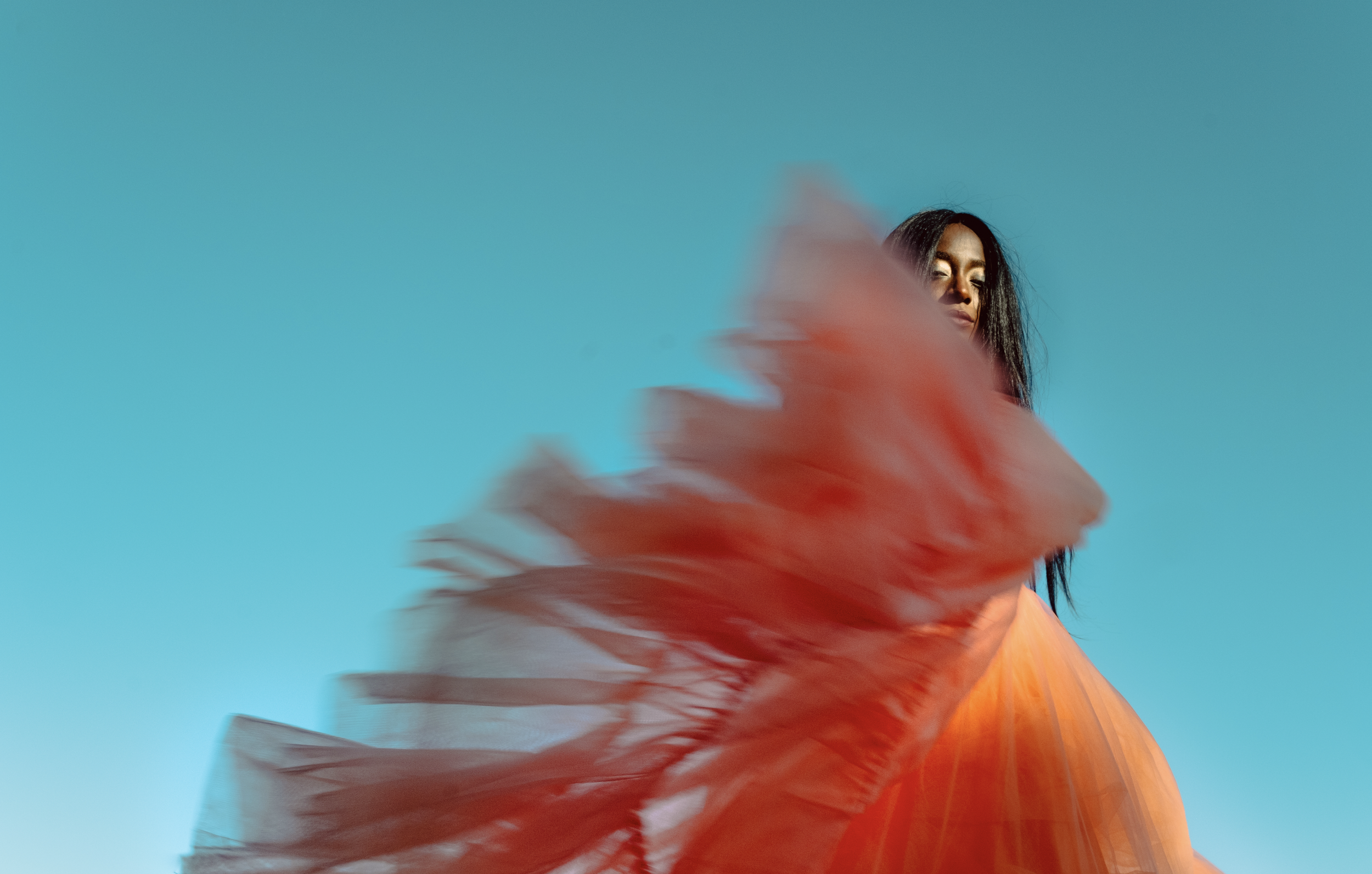 A woman with long dark hair standing against a clear blue sky, wearing an orange flowing dress with a red fabric swirling around her.