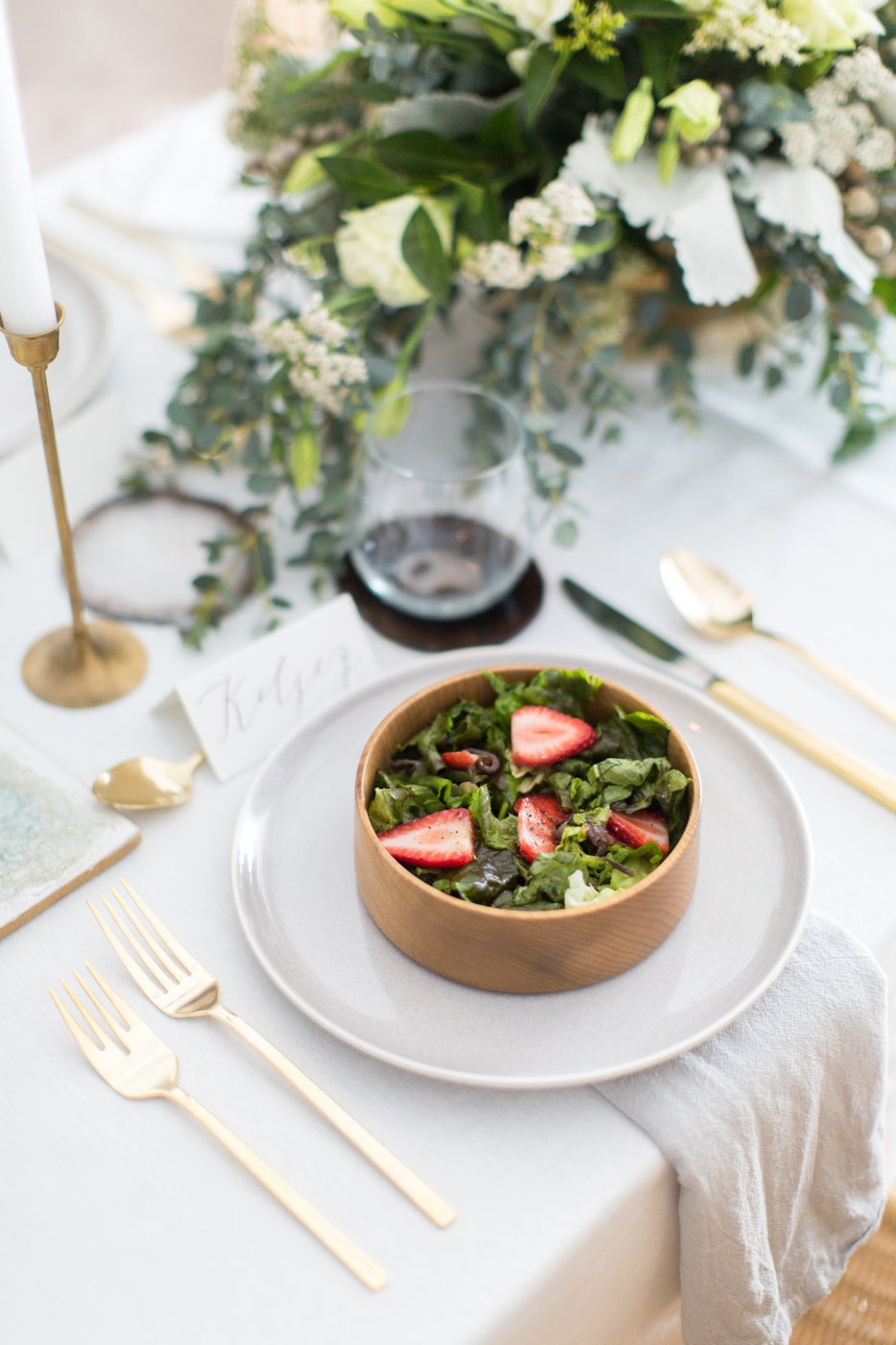 A table setting featuring a bowl of salad with strawberries, gold utensils, a glass of red wine, a candle holder, and a floral centerpiece with white and green flowers.