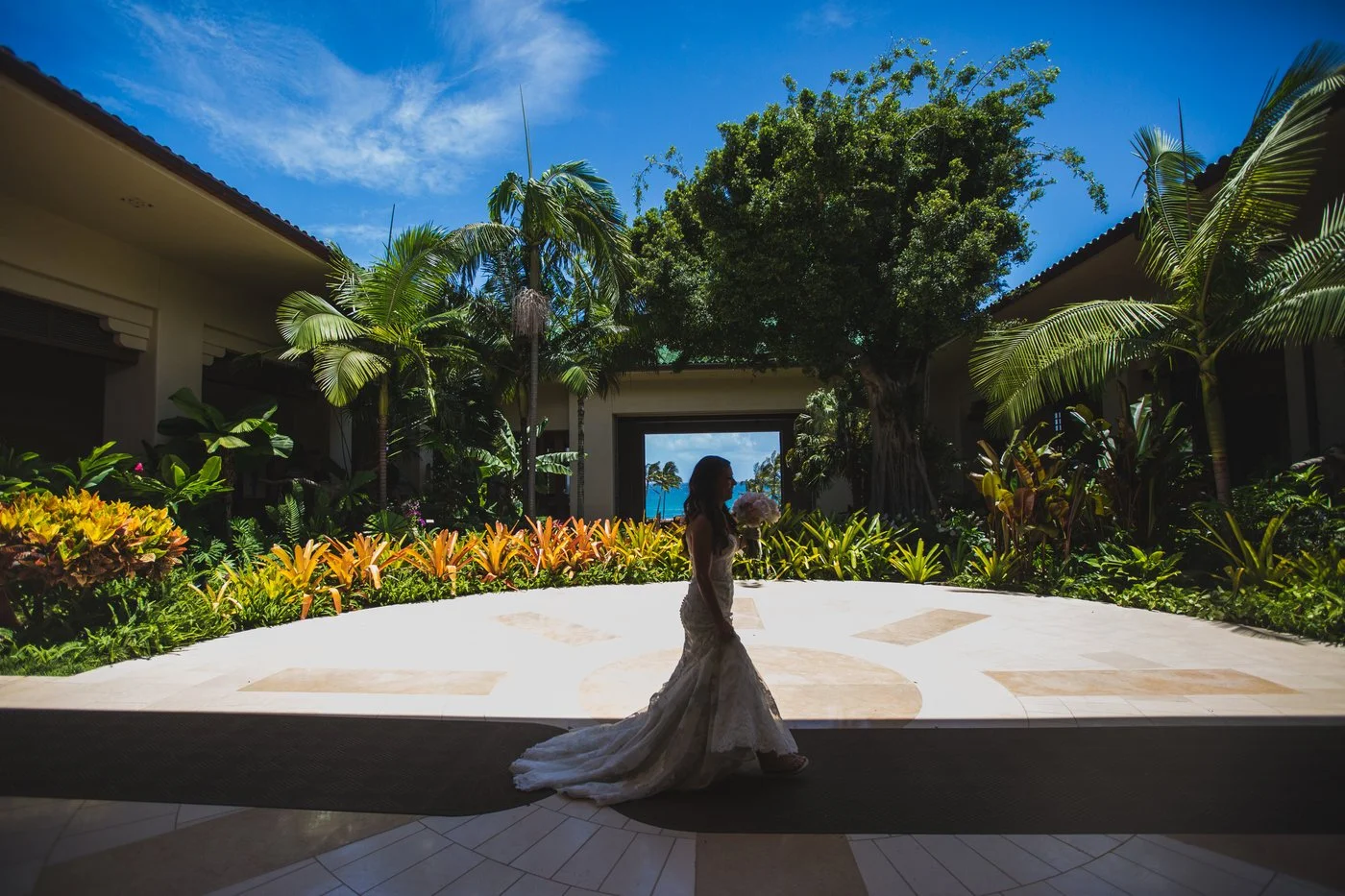 A bride in a wedding dress holding a bouquet, silhouetted against a bright outdoor background with tropical plants, palm trees, and a view of the ocean through an opening.