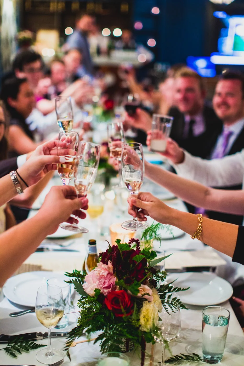 People raising glasses in a toast at a formal dinner party with a floral centerpiece on the table.