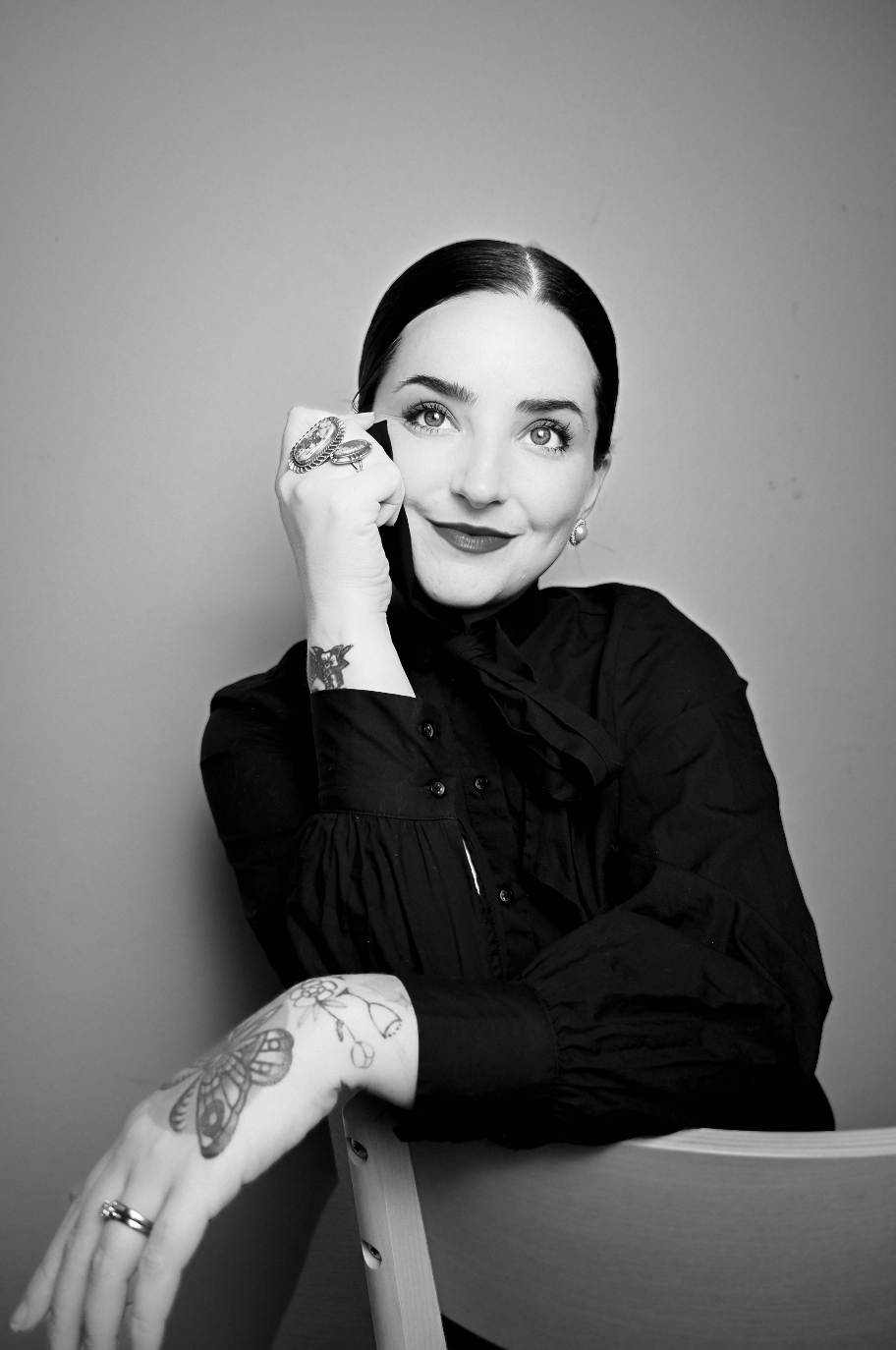 Black and white portrait of a woman with dark hair, wearing a dark blouse, smiling and holding a phone to her ear. She has tattoos on her left arm, rings on her fingers, and earrings. She is sitting against a plain background.