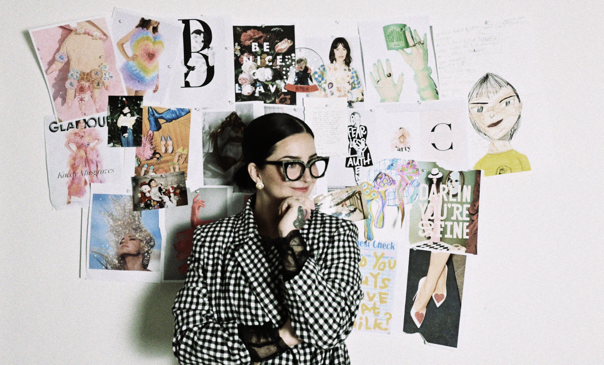 A woman with dark hair in a bun, wearing large black glasses and a black and white checkered blazer, stands pensively in front of a wall decorated with various colorful pictures, illustrations, and posters.