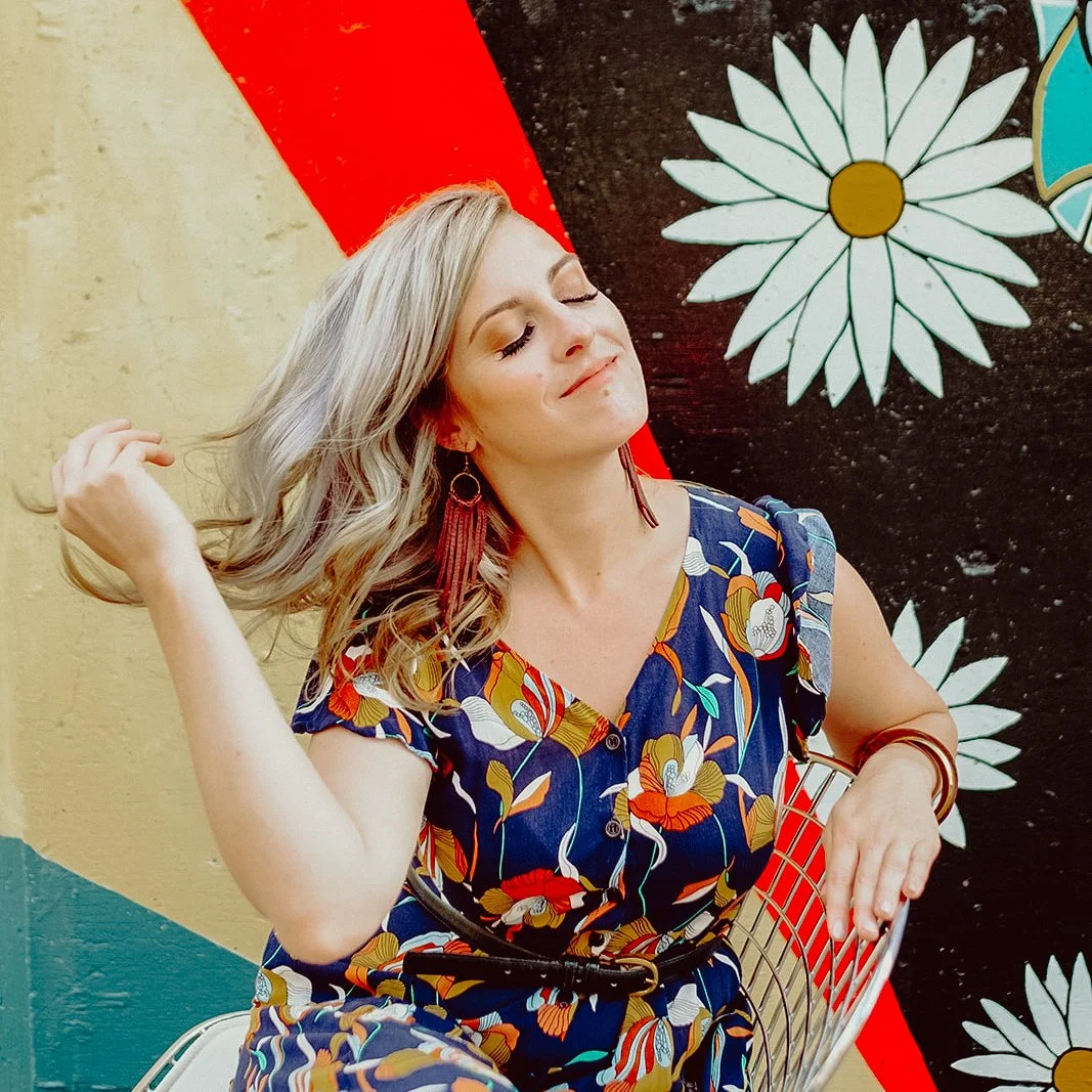 A woman with blonde hair and closed eyes smiling, wearing a colorful floral dress and large earrings, sitting on a white chair against a flower-themed mural background.
