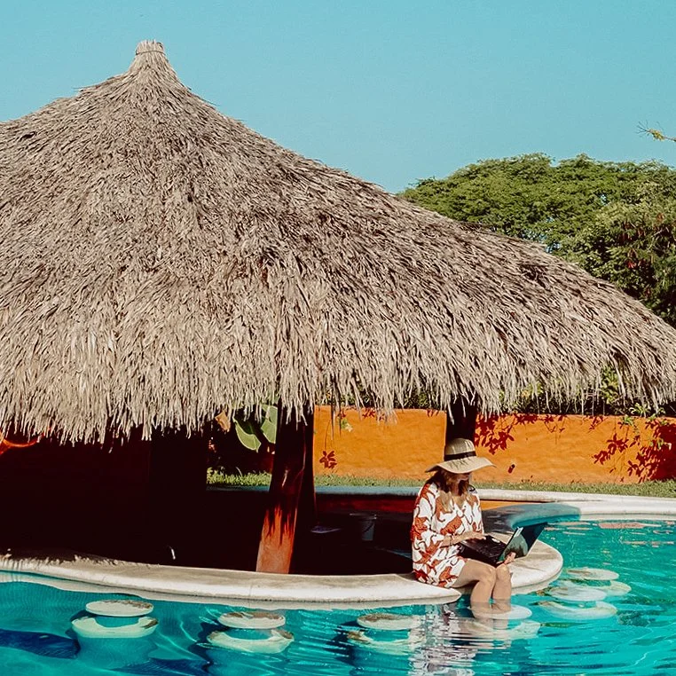 Woman sitting by the pool under a thatched umbrella, using a laptop, wearing a large sun hat and a floral cover-up.