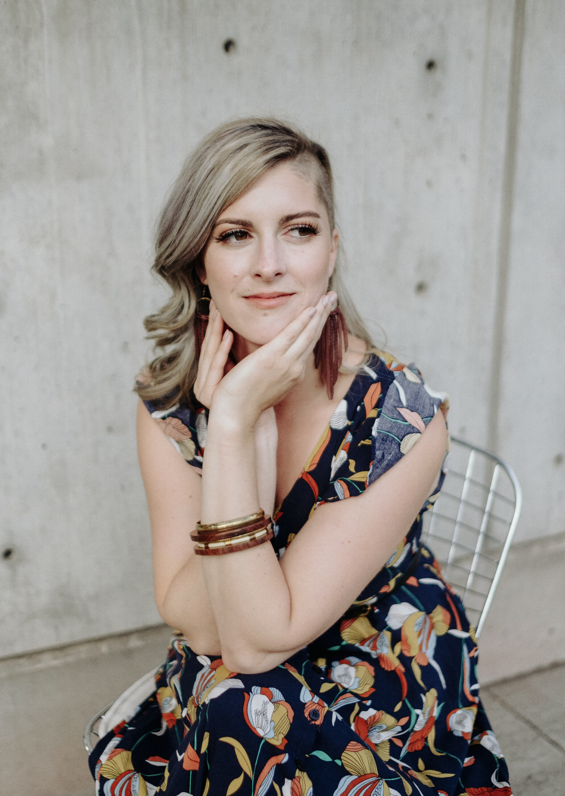 A woman with short, wavy blonde hair and light skin, wearing a colorful floral dress and brown bracelets, sits on a wire chair outdoors, resting her chin on her hand and looking thoughtfully to the side against a plain concrete wall background.