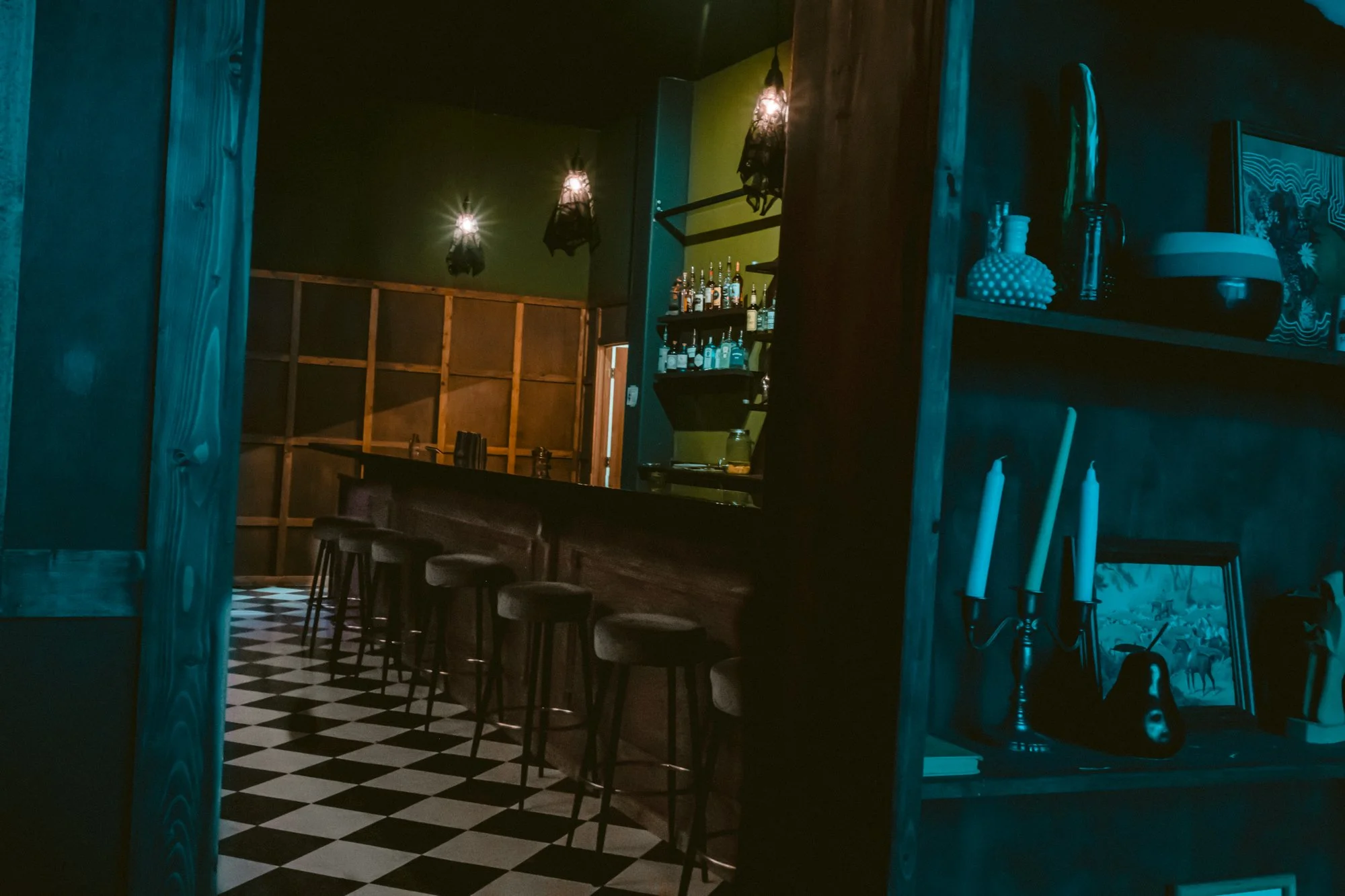 Interior of a dimly lit bar with a checkered black-and-white floor, a row of barstools along a wooden counter, and shelves with bottles and decorative items.