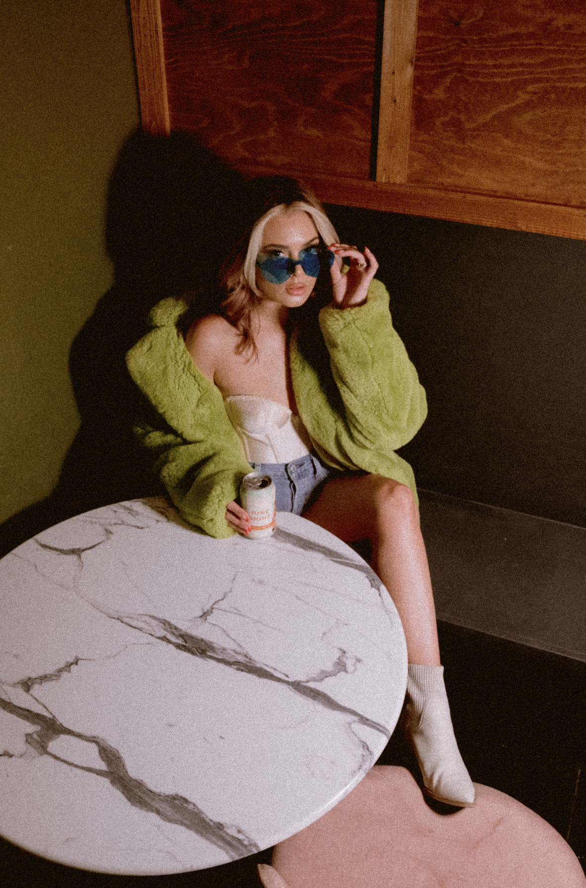 A woman with blonde and pink hair sitting on a black bench against a wood-paneled wall, wearing a green fuzzy jacket, white corset top, denim shorts, and white boots, holding a can of soda and adjusting blue sunglasses.