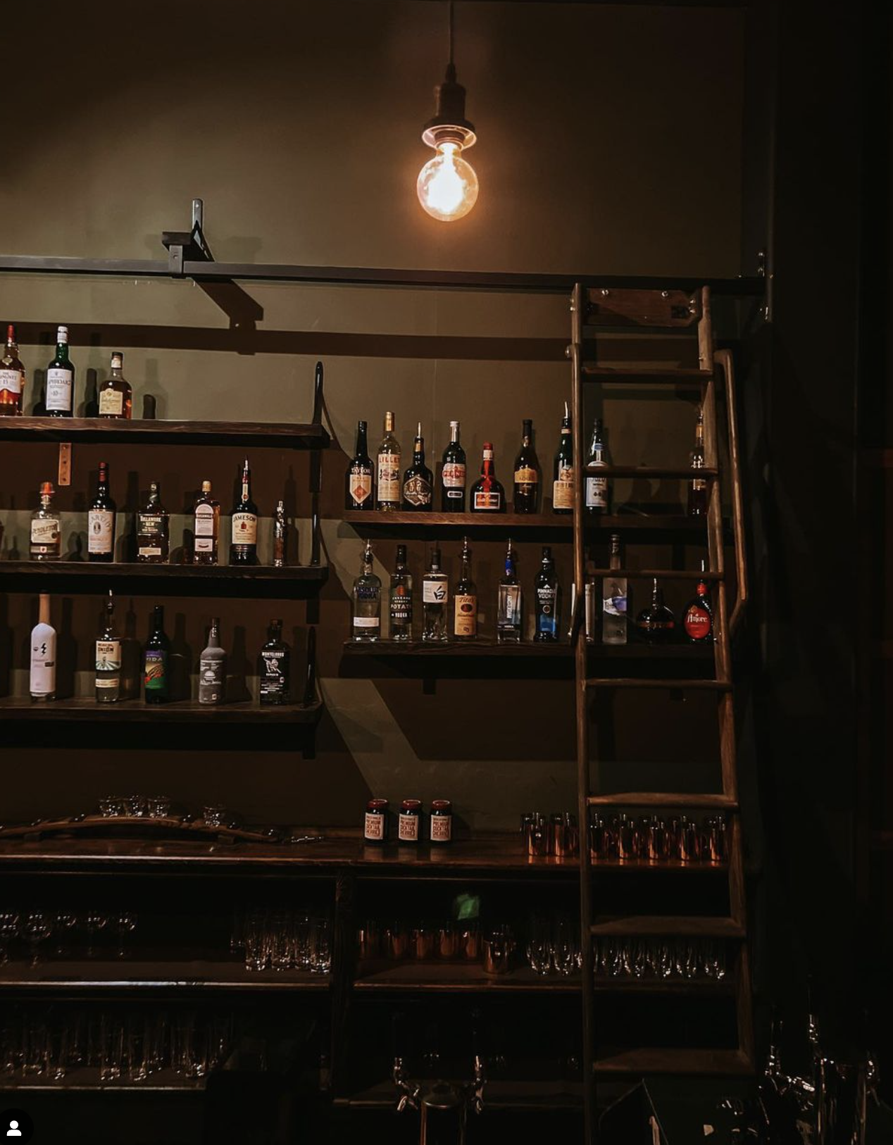 A warm-lit bar scene with shelves of various liquor bottles, a wooden ladder leaning against the wall, and an exposed light bulb hanging from the ceiling, creating a cozy atmosphere.