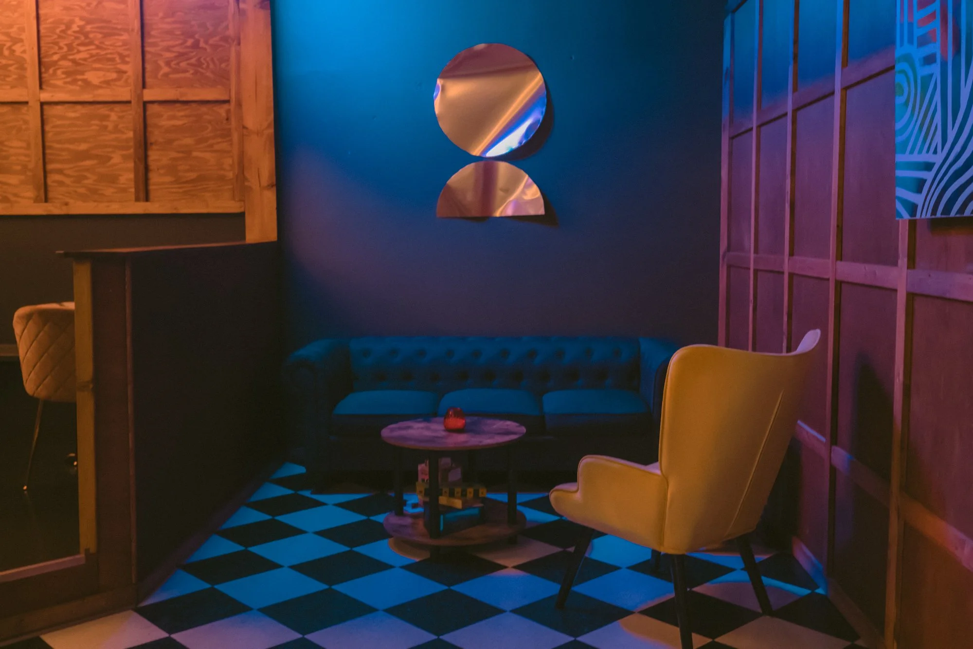 Dimly lit lounge with a tufted blue sofa, yellow armchair, and a small round coffee table, against a blue wall with metallic wall art. Checkerboard floor adds to retro ambiance.