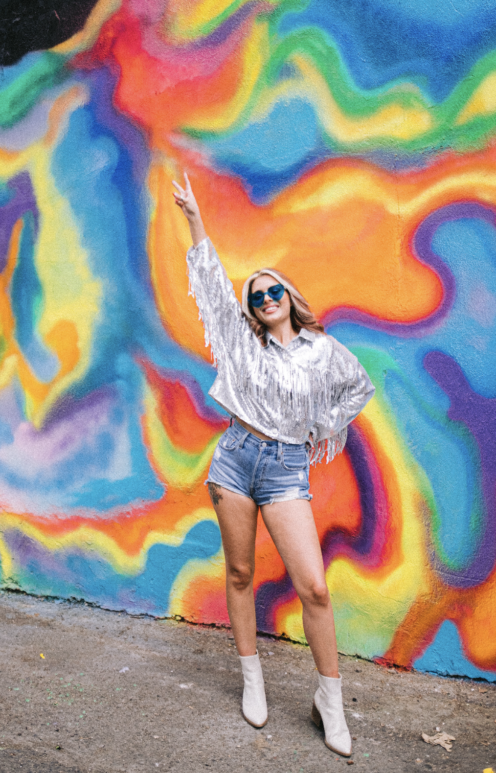 Young woman wearing sunglasses, a shiny silver shirt, denim shorts, and white boots, standing with one arm raised and making a peace sign, in front of a colorful abstract mural wall.