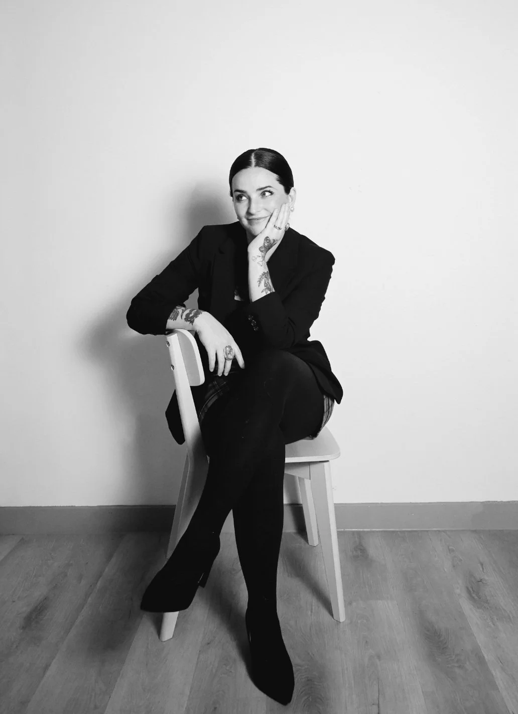 A young woman with dark hair, tattoos on arms, wearing a black blazer and black pants, sitting on a chair against a plain white wall, smiling and looking to her left.