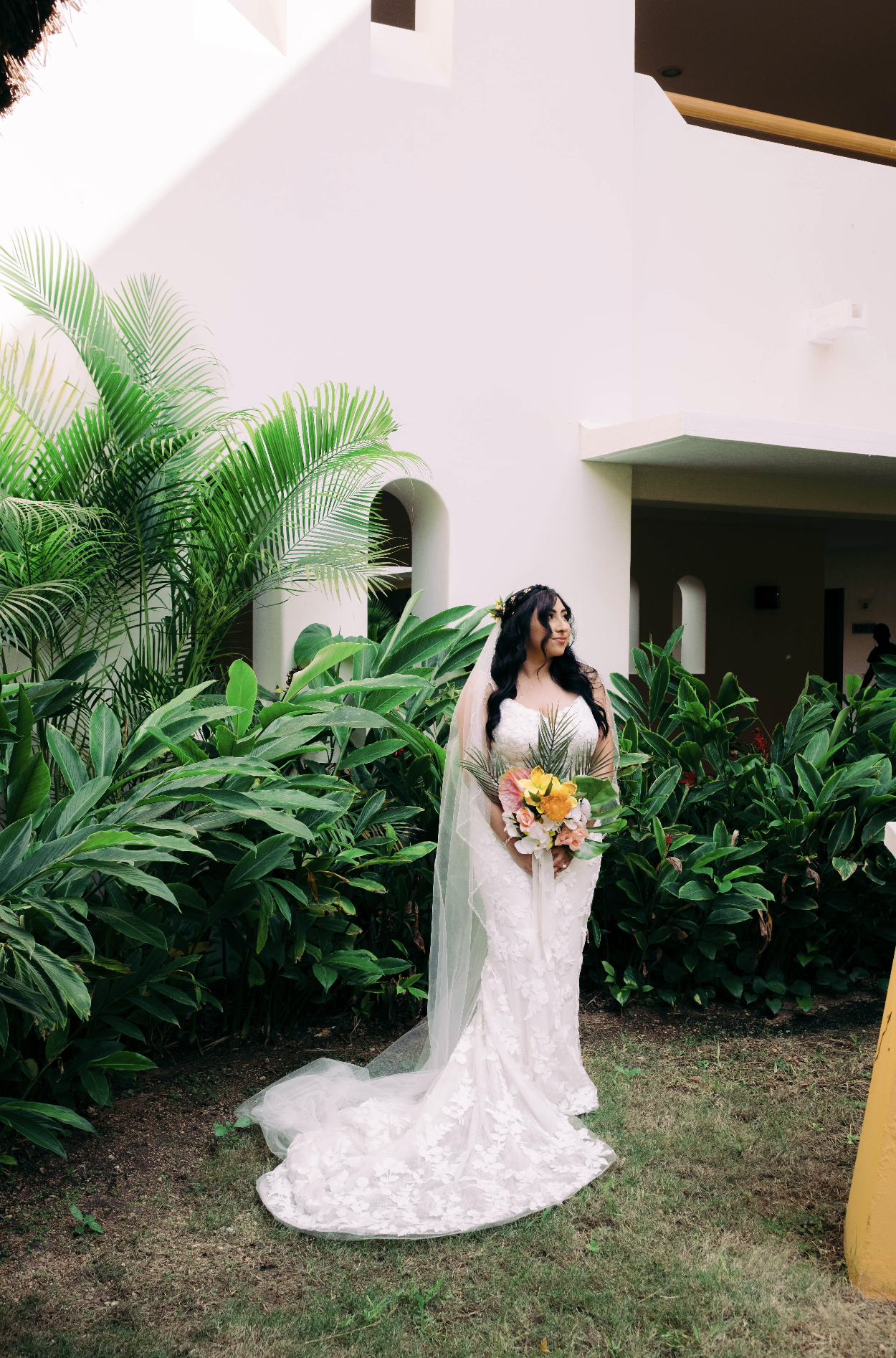 Bride in a white lace wedding dress and veil holds a bouquet of yellow and pink flowers, standing among green tropical plants in front of a white building with curved archways.