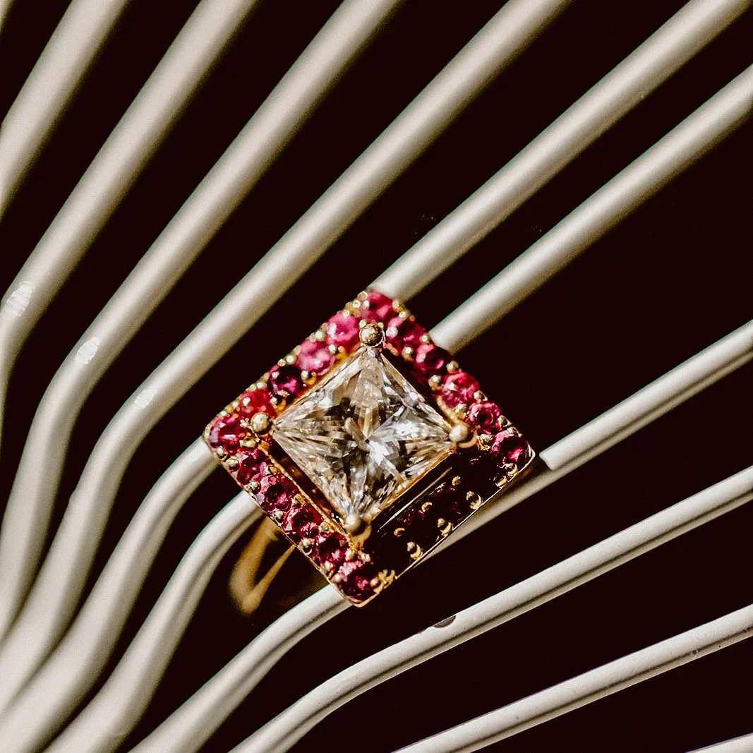 A ring with a large square-shaped central diamond surrounded by small pink and red gemstones, resting on a silver ribbed surface.