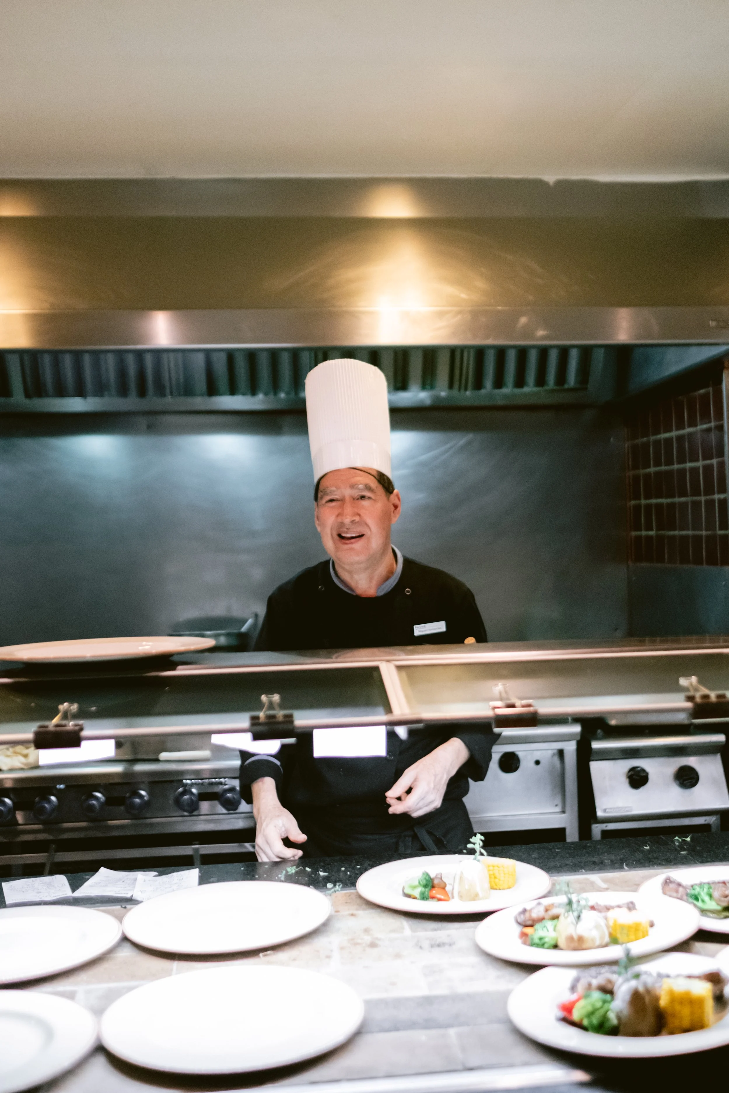 A chef in a white chef hat and black uniform laughs behind a kitchen counter with plated dishes, with a stainless steel kitchen hood above him.