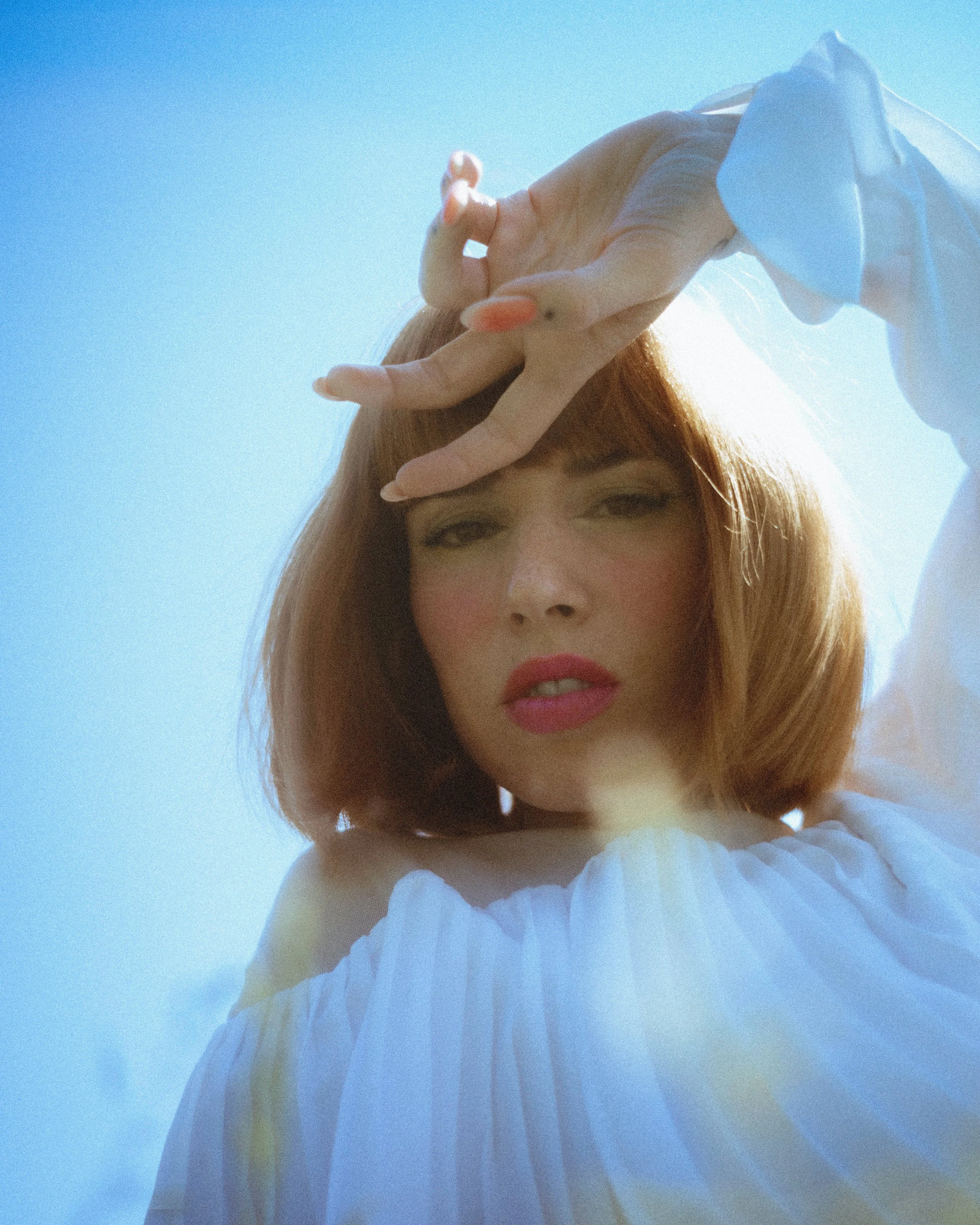 A woman with reddish hair, wearing a white off-shoulder top, is looking at the camera with a hand near her face that has a turtle-shaped nail polish. She is outdoors with a clear blue sky in the background.