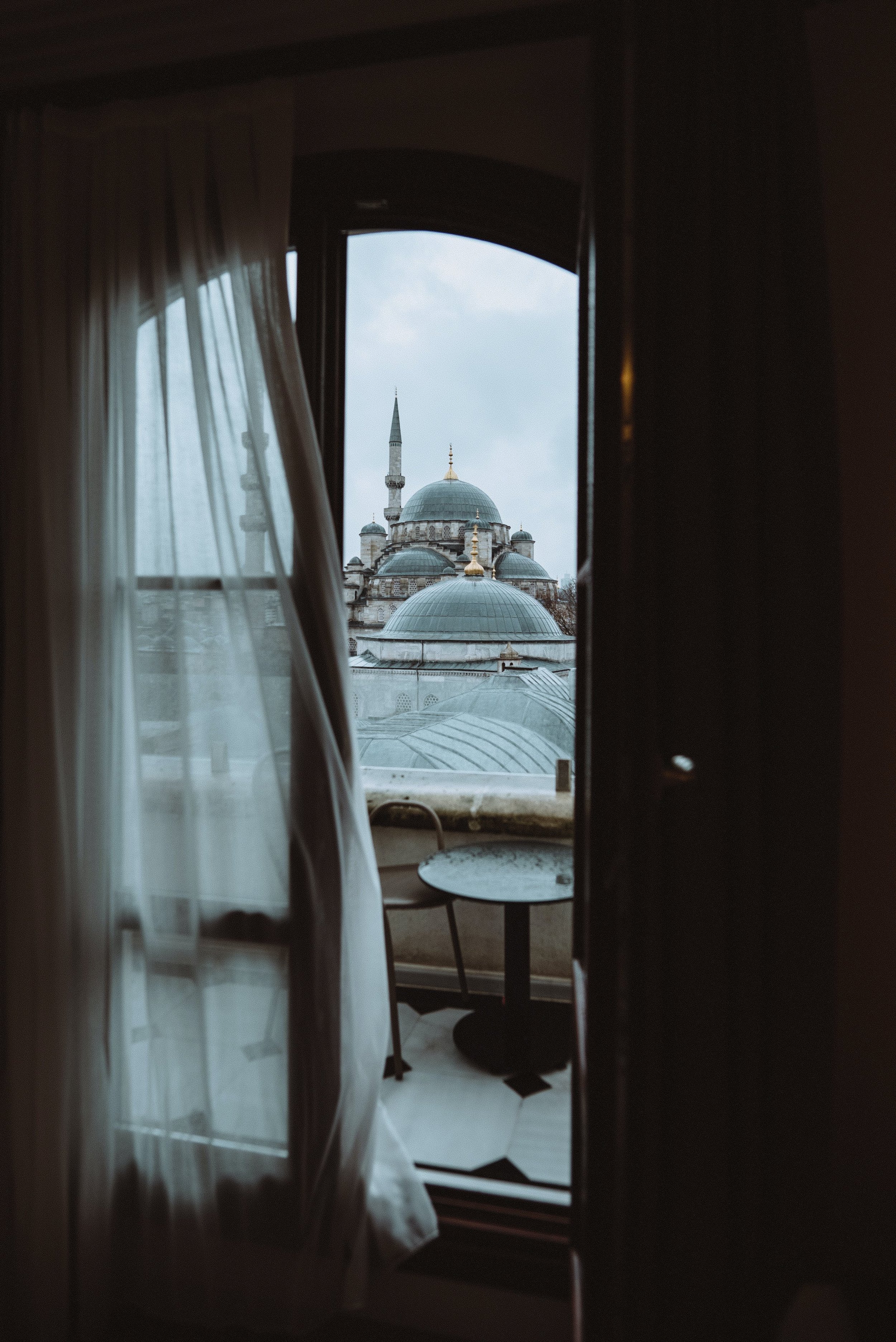 View of a mosque through a partially open window with curtains, showing domes and a minaret.
