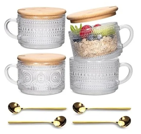 Four glass storage containers with wooden lids, one filled with cereal and fruit, and gold-colored spoons in front.