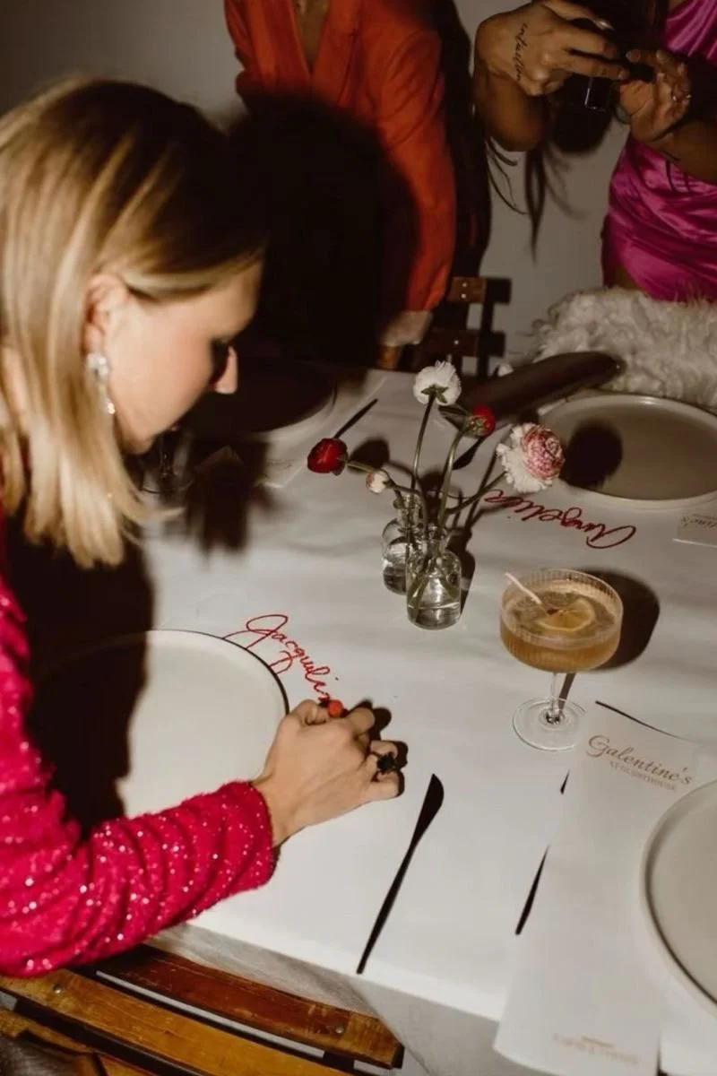 A woman with blonde hair in a red sparkly dress signs a large white placemat at a table decorated for a celebration, with a cocktail and small vases of flowers nearby.