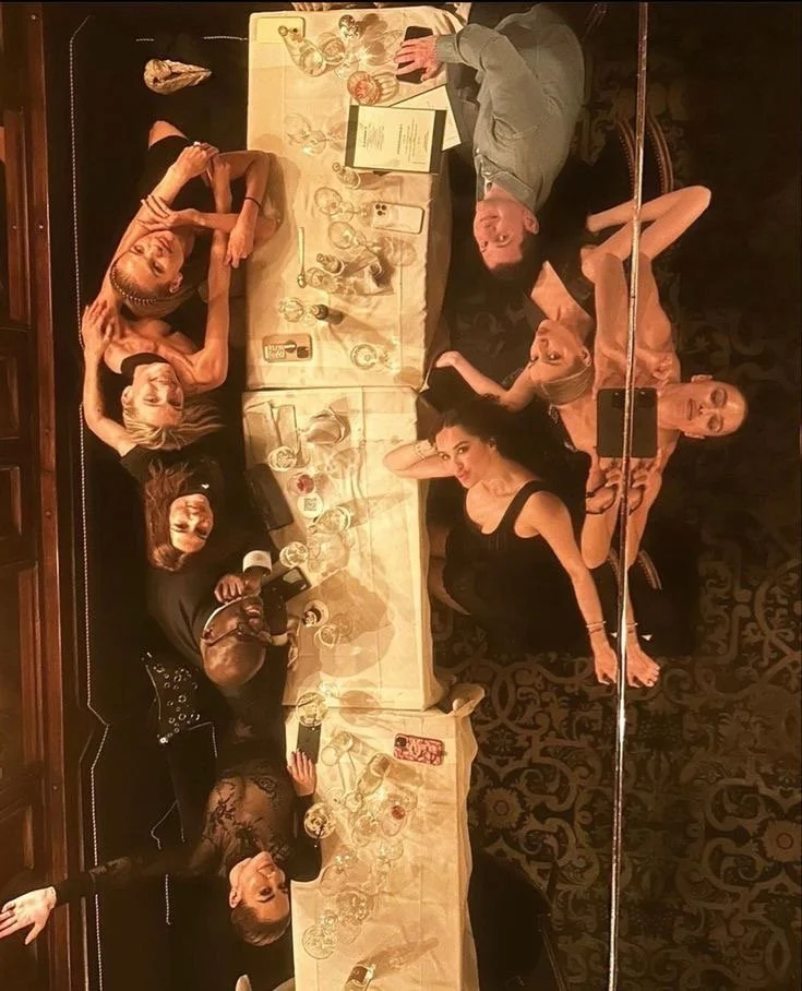 Group of six people, five women and one man, lying down at a table with multiple glasses, bottles, and a menu. The photo is taken from above, and they are posing for the camera with relaxed expressions, in what appears to be a restaurant or banquet setting.