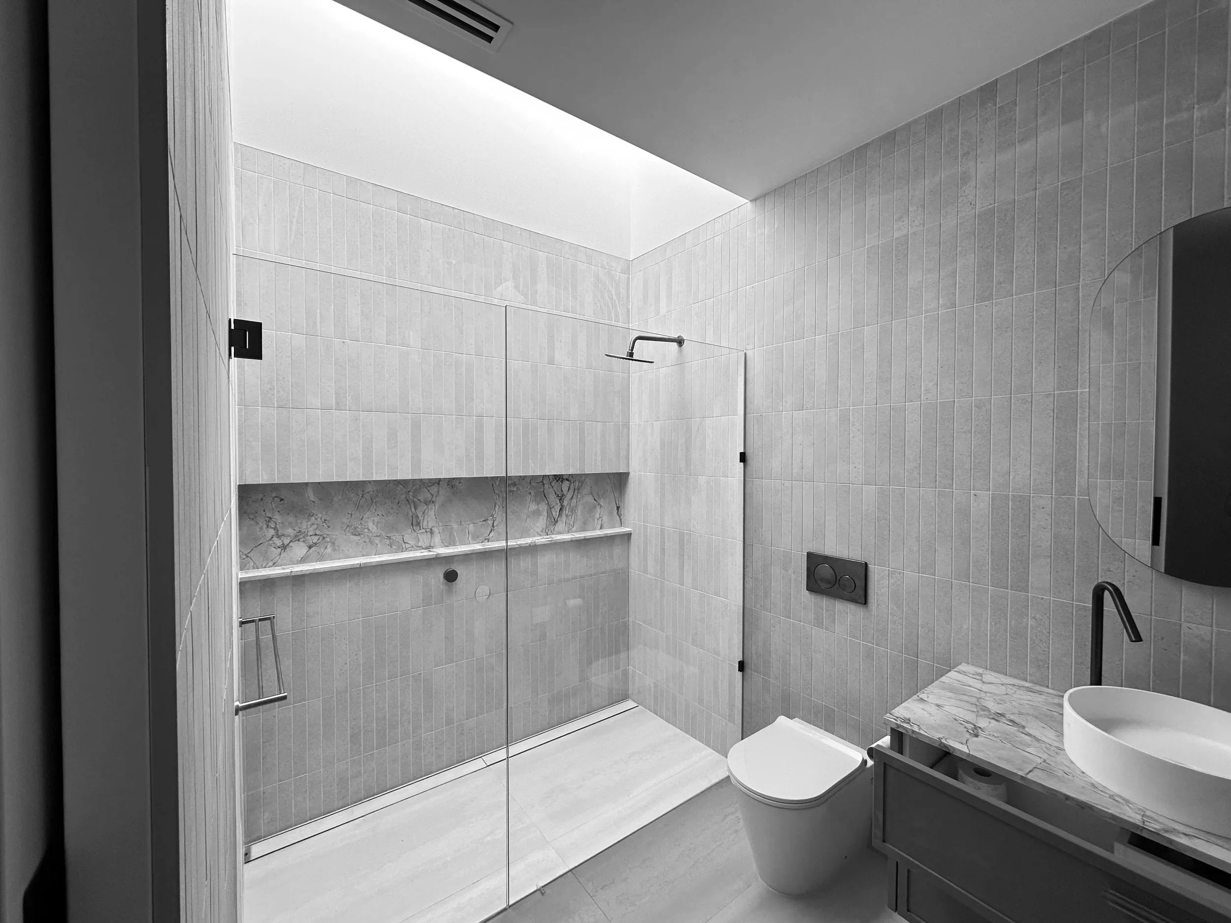 Modern minimalist bathroom with glass shower enclosure, marble ledge, wall-mounted toilet, and marble countertop sink with black faucet.