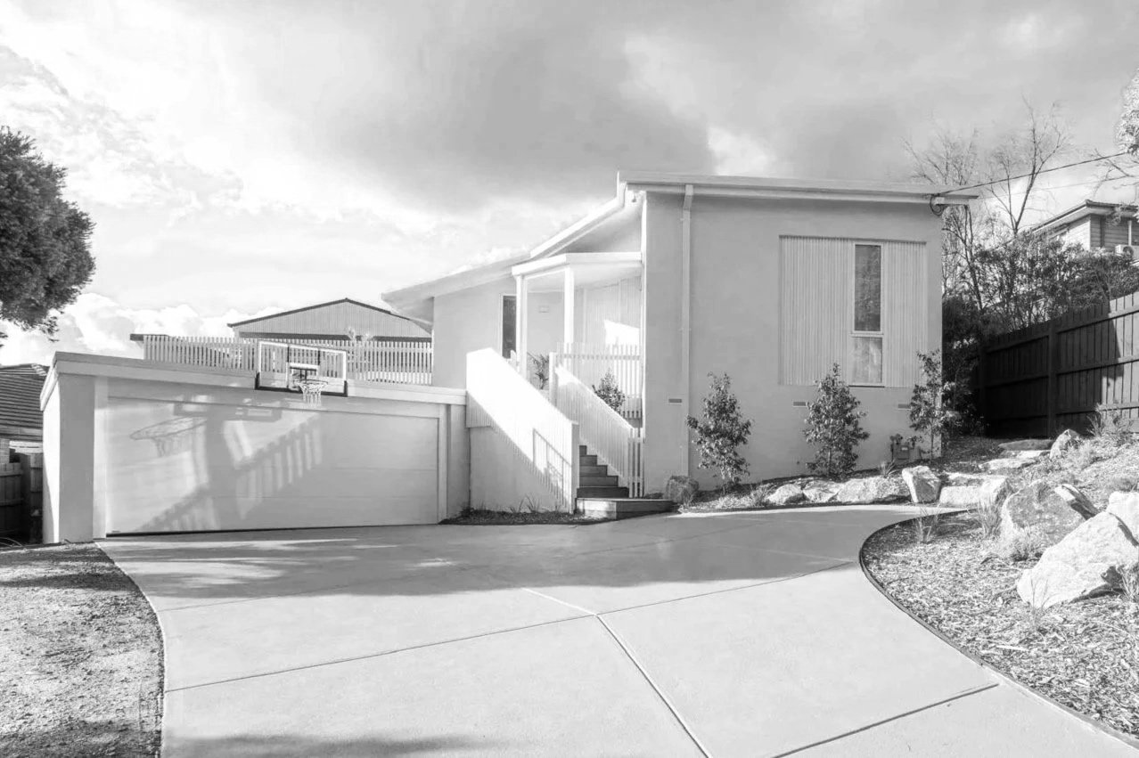 A modern two-story house with a driveway, a garage, steps leading up to a front porch, and a small basketball hoop above the garage door, surrounded by trees and rocks.