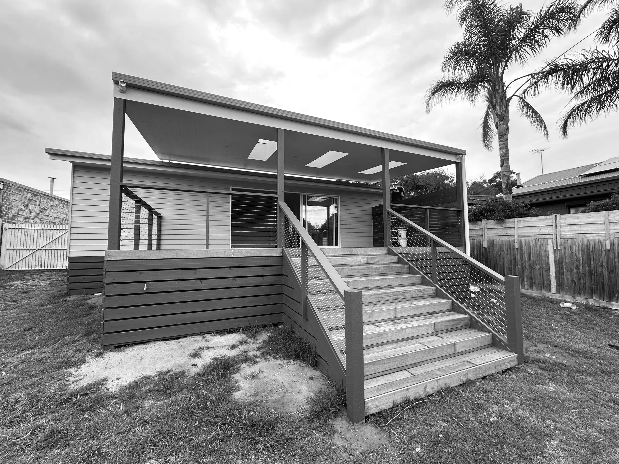 Backyard view of a house with a wooden deck, stairs, and a covered roof, surrounded by a grassy area and a wooden fence, with palm trees and neighboring houses in the background.
