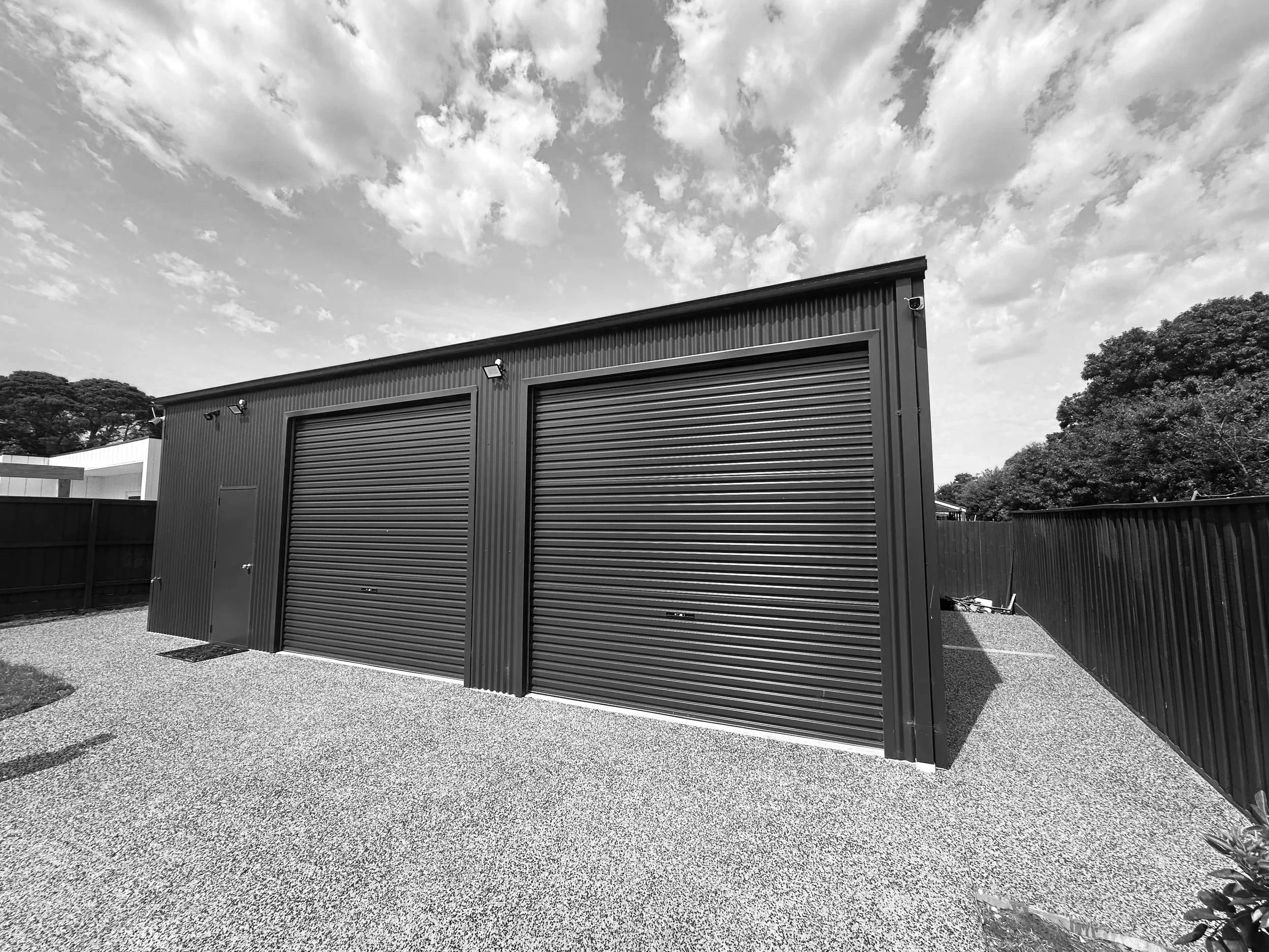 A black metal building with two large roll-up doors and one side door, situated in a gravel yard enclosed by a black fence, with a partly cloudy sky overhead and trees in the background.