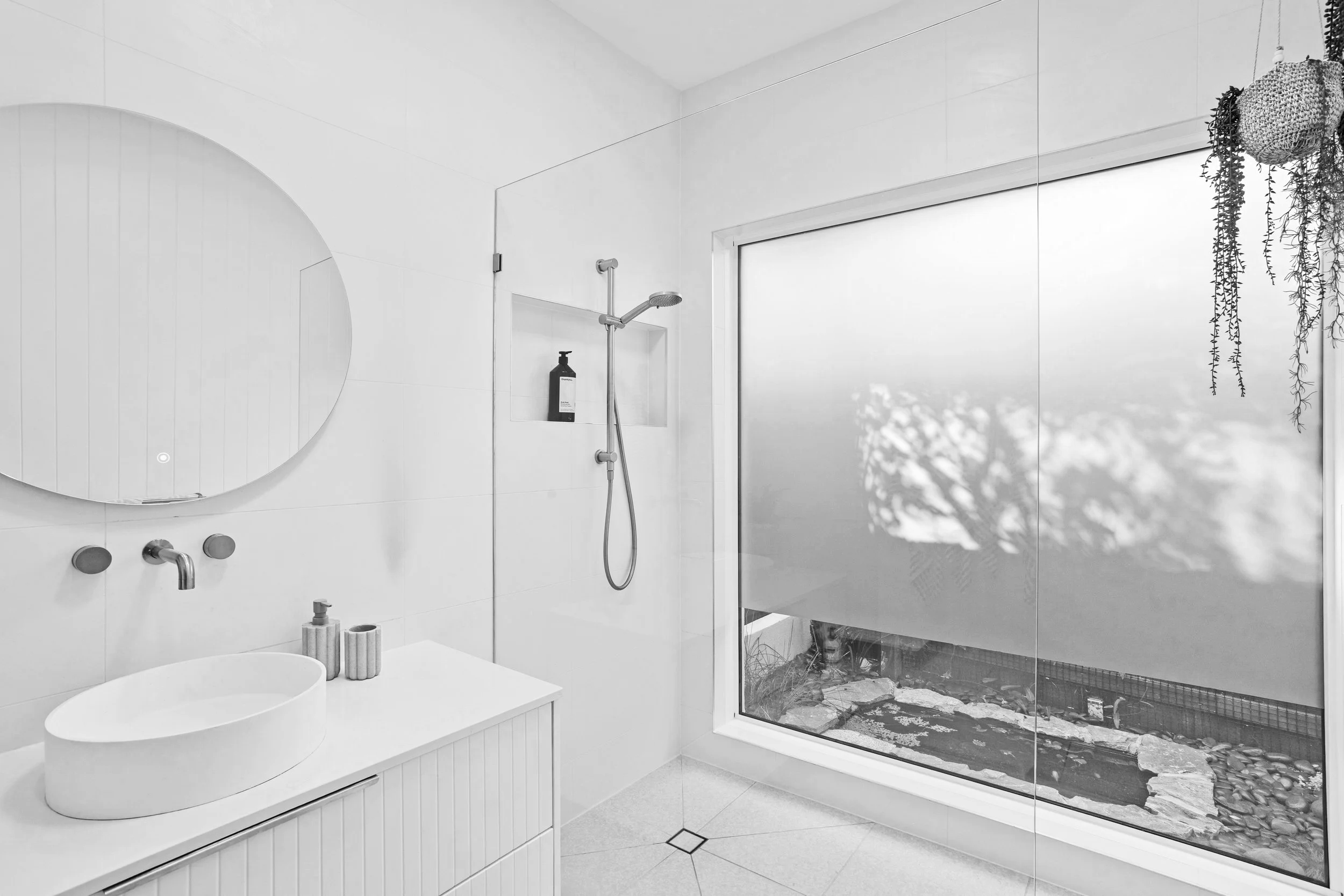 A modern, minimalist bathroom with a white vanity, oval sink, circular mirror, and a large frosted window. There is a shower head with a bottle on a built-in shelf and some hanging plants in the corner.