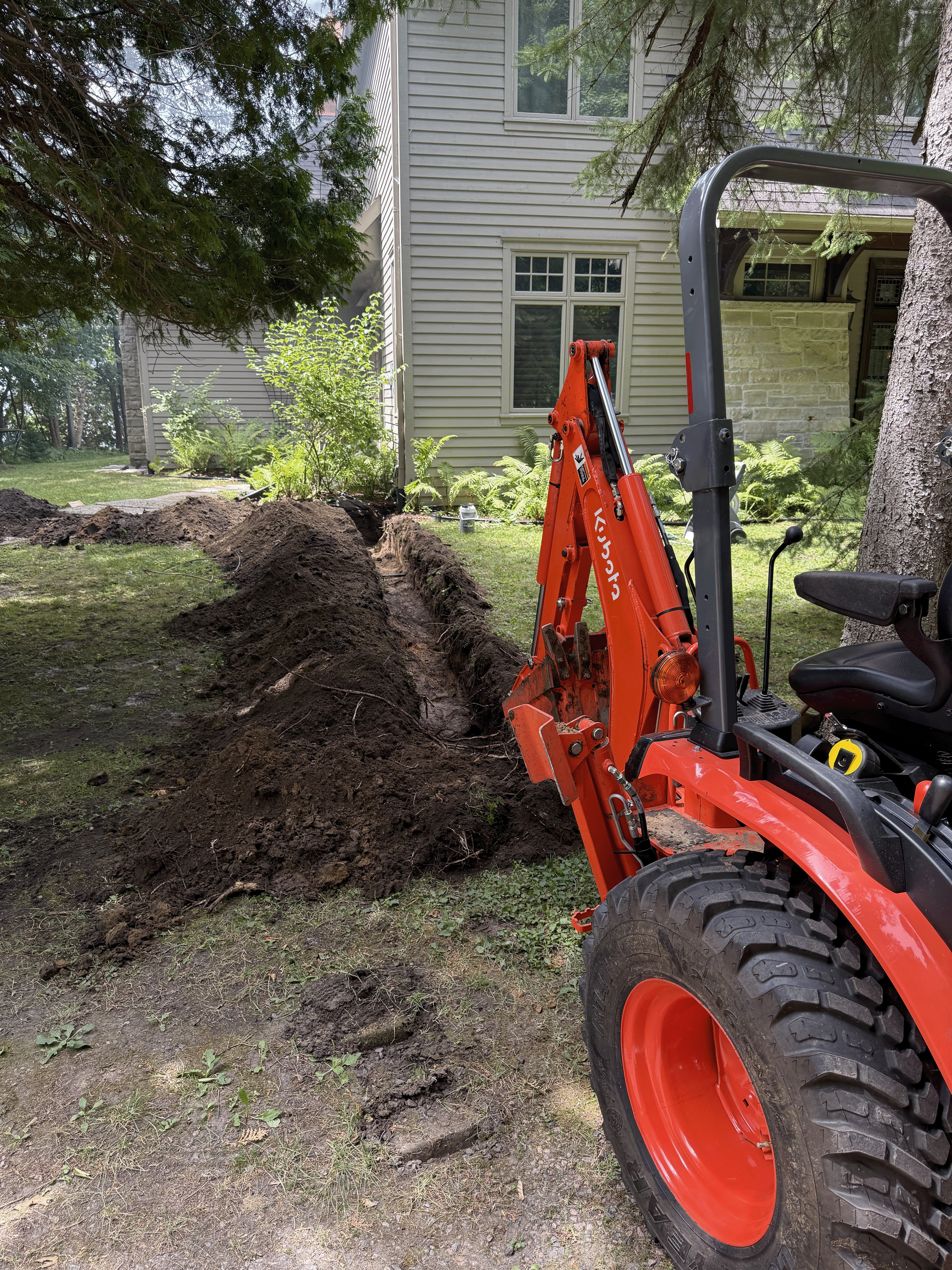 A small orange Kubota tractor in the process of digging a trench in a backyard with a house in the background, surrounded by trees and bushes.