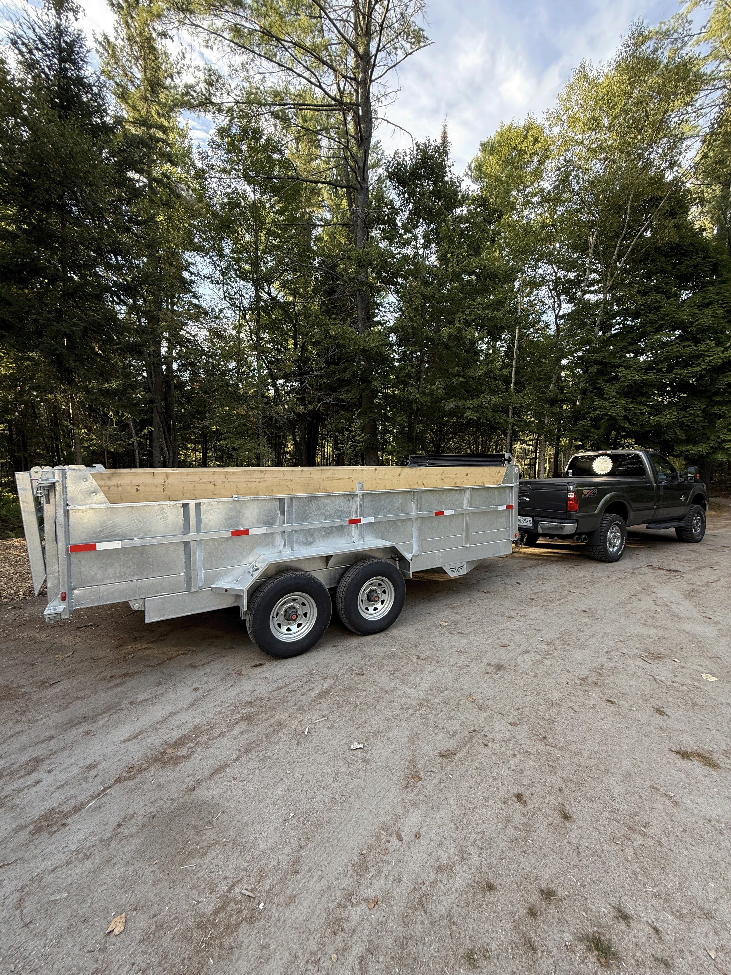 Black pickup truck attached to a silver utility trailer with wooden sides, parked on a dirt surface with trees in the background.